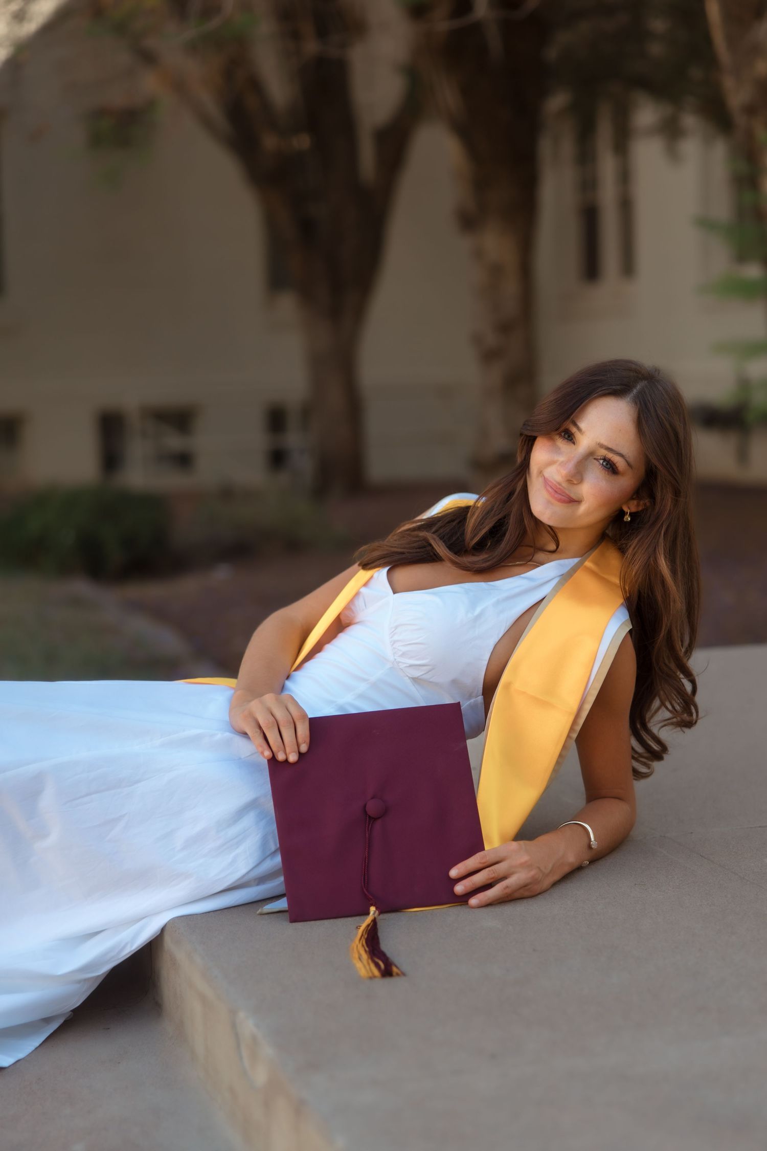 Young woman in white graduation gown with gold sash holds maroon cap while posing outdoors on campus steps.