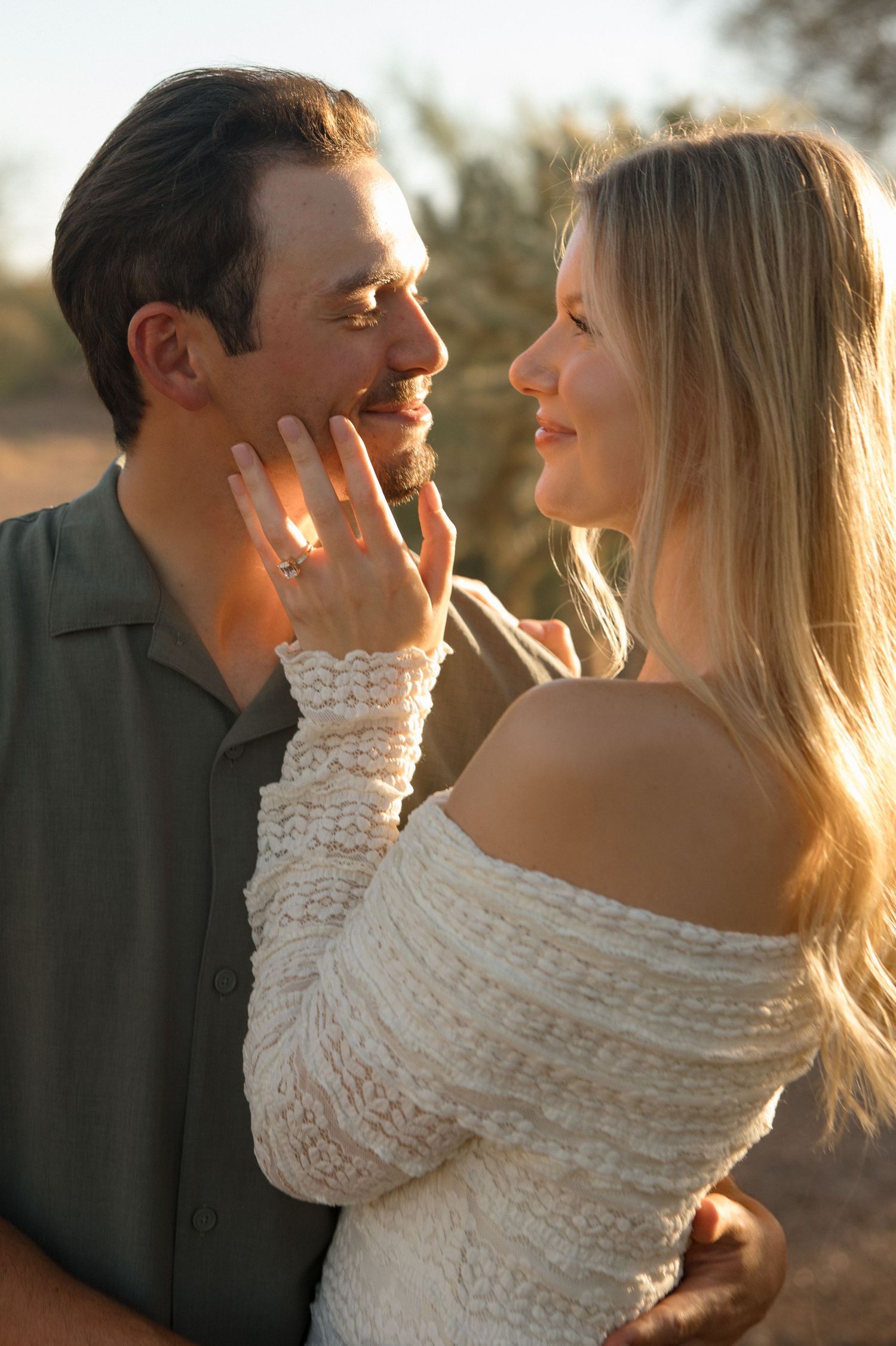 Romantic couple sharing a tender moment outdoors at golden hour, woman in white lace top touching man's face.
