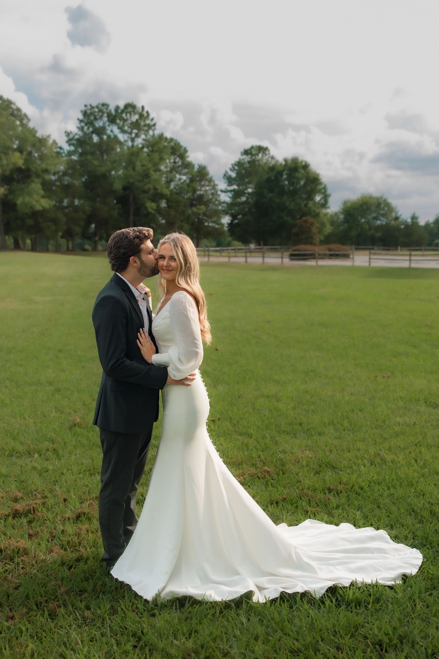 Bride in white gown with train and groom in navy suit share a kiss in a lush green field.