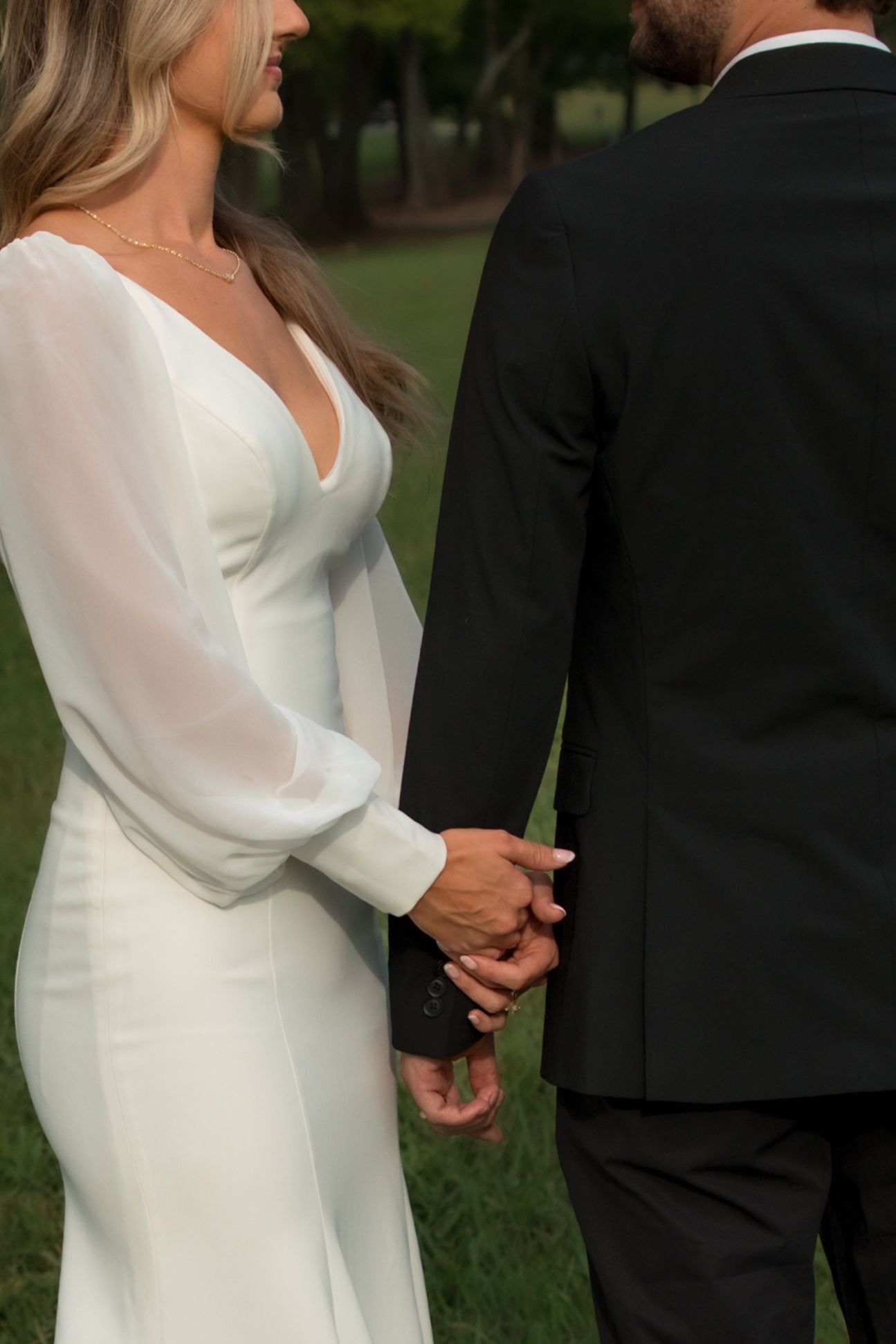 Bride in white long-sleeve gown holding hands with groom in black suit outdoors on green grass.