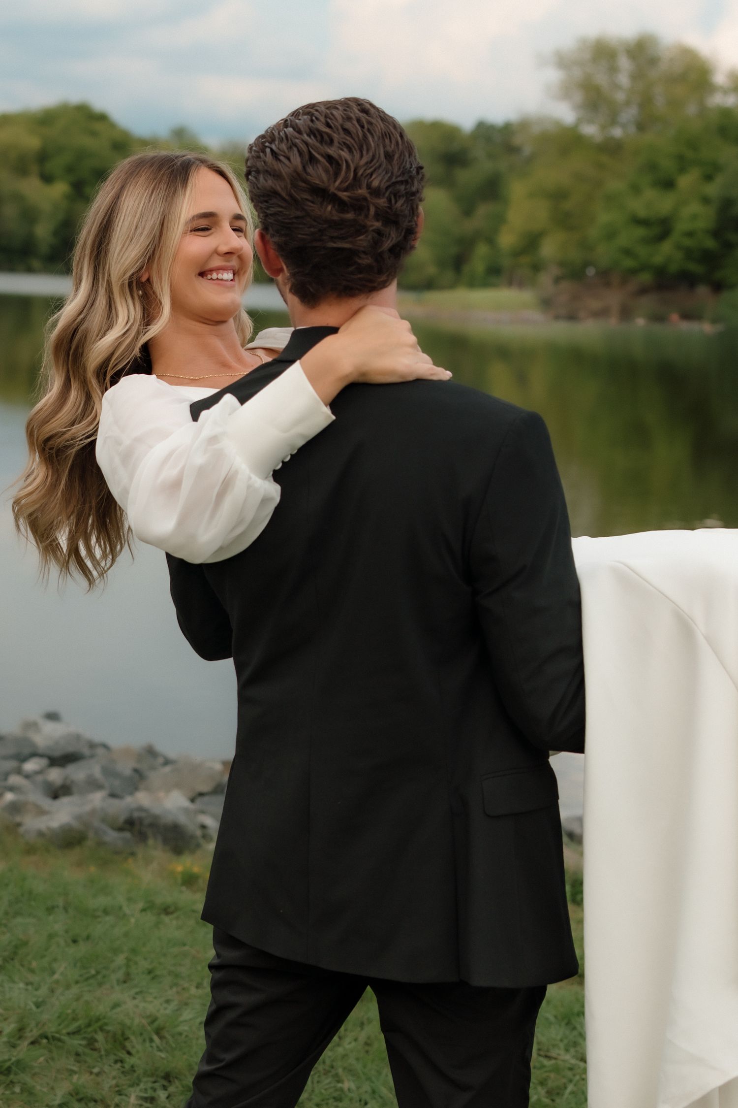 Smiling bride with flowing hair embraces groom in black suit by a serene lake surrounded by trees.