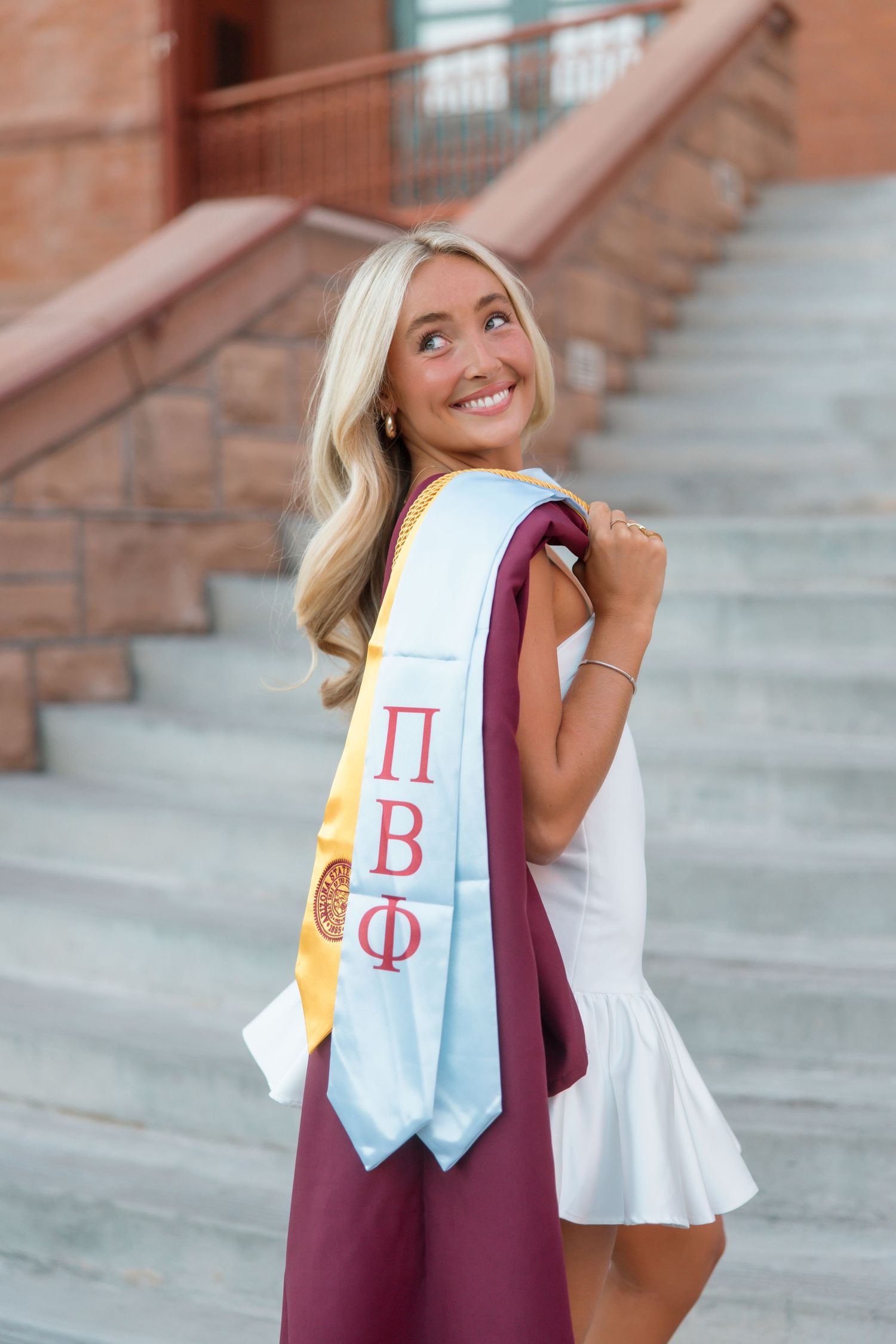 Smiling blonde graduate wearing white dress with Pi Beta Phi sash and maroon graduation gown on university steps.