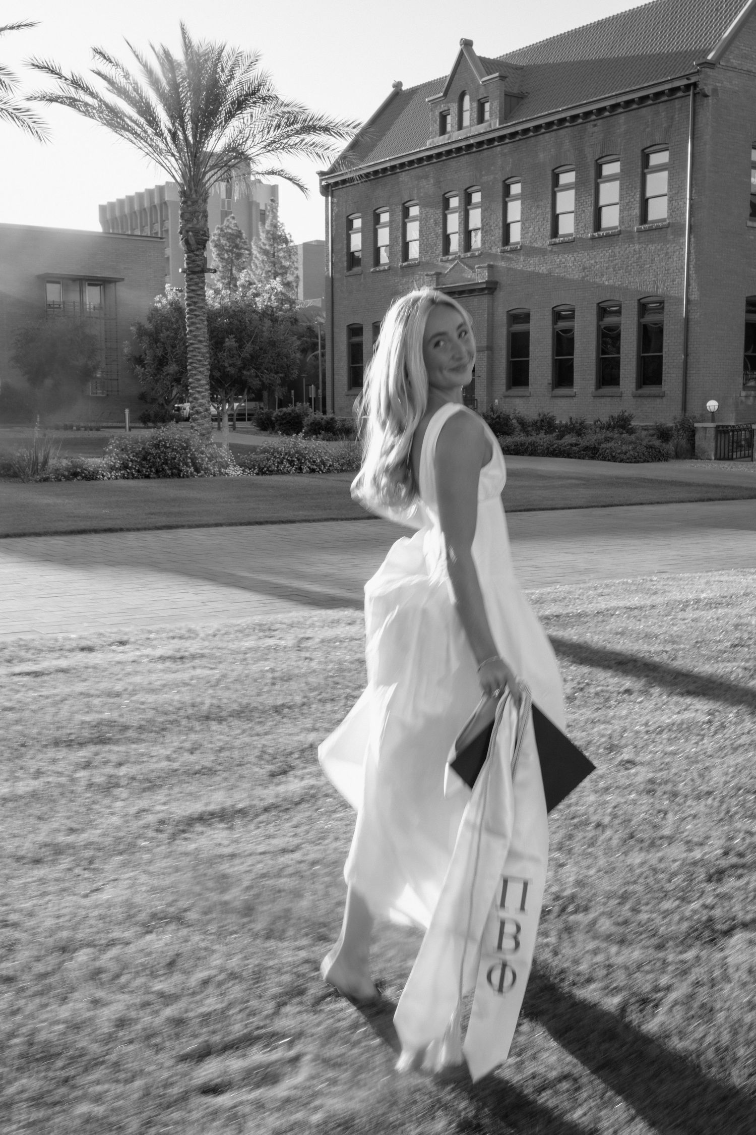Black and white photo of blonde graduate in white dress with Pi Beta Phi stole on sunny campus.