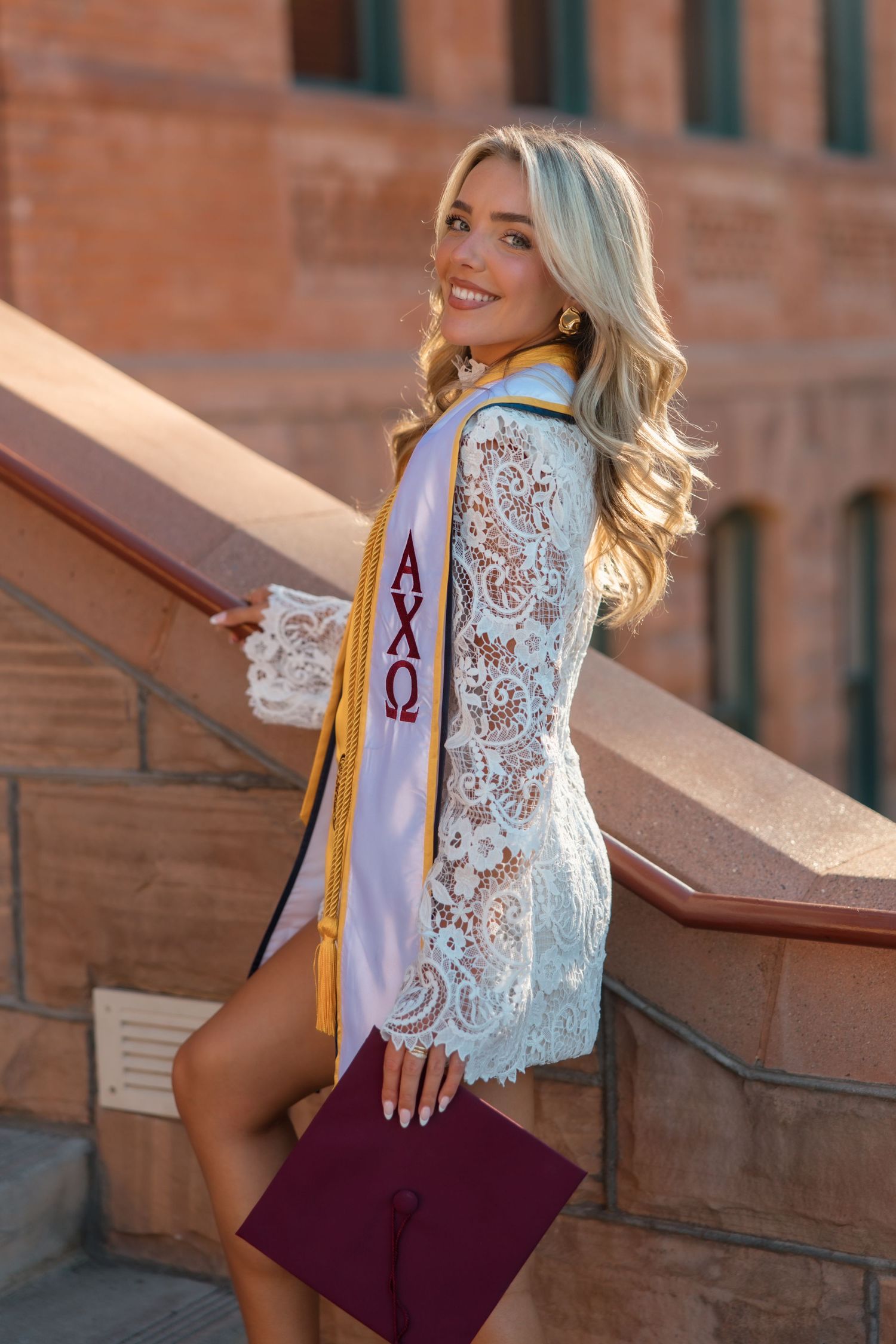 Smiling blonde graduate poses on stone steps wearing lace dress and Alpha Chi Omega sash holding maroon cap.