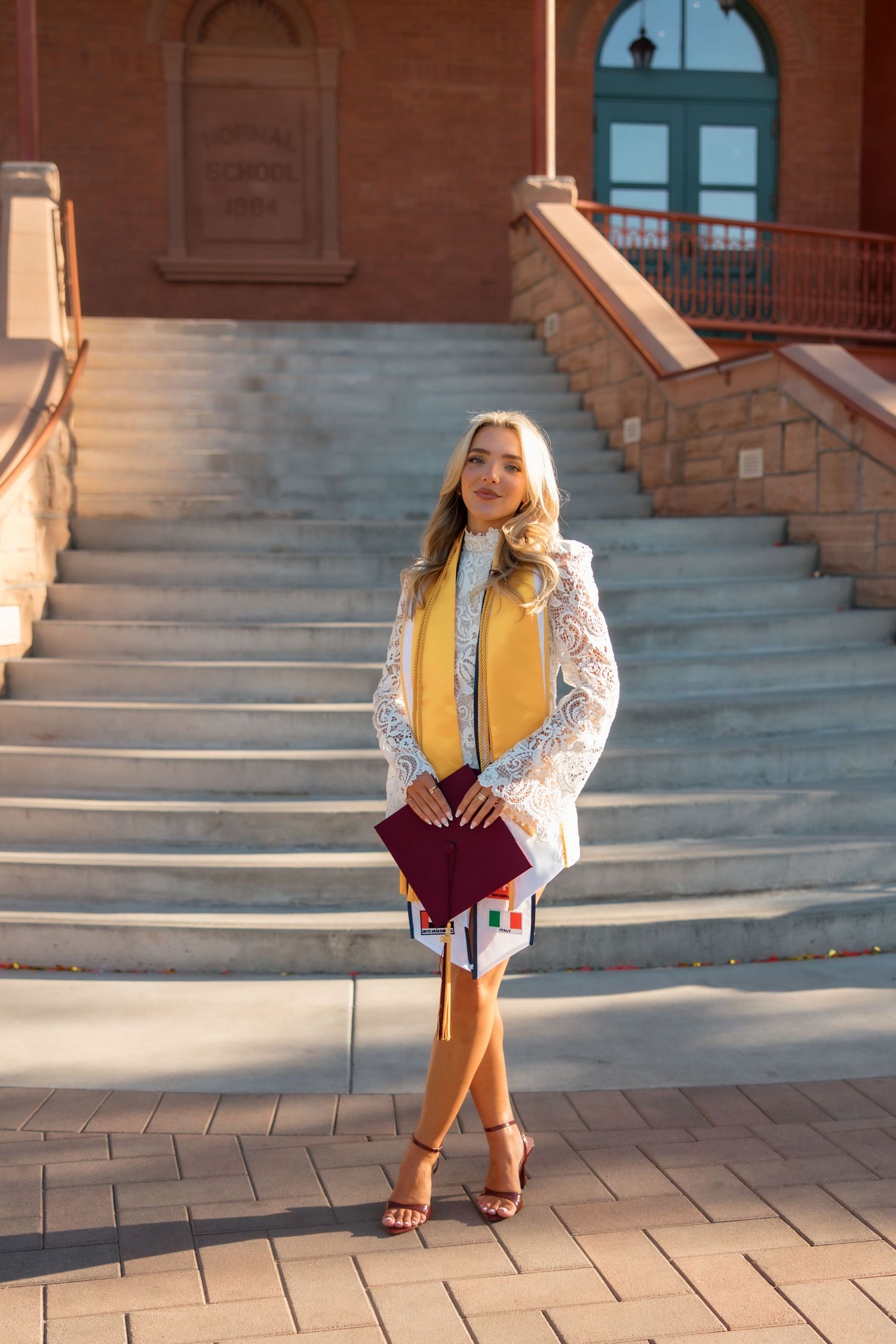 Young woman in white lace jacket and yellow graduation stole holds diploma cover on university steps.