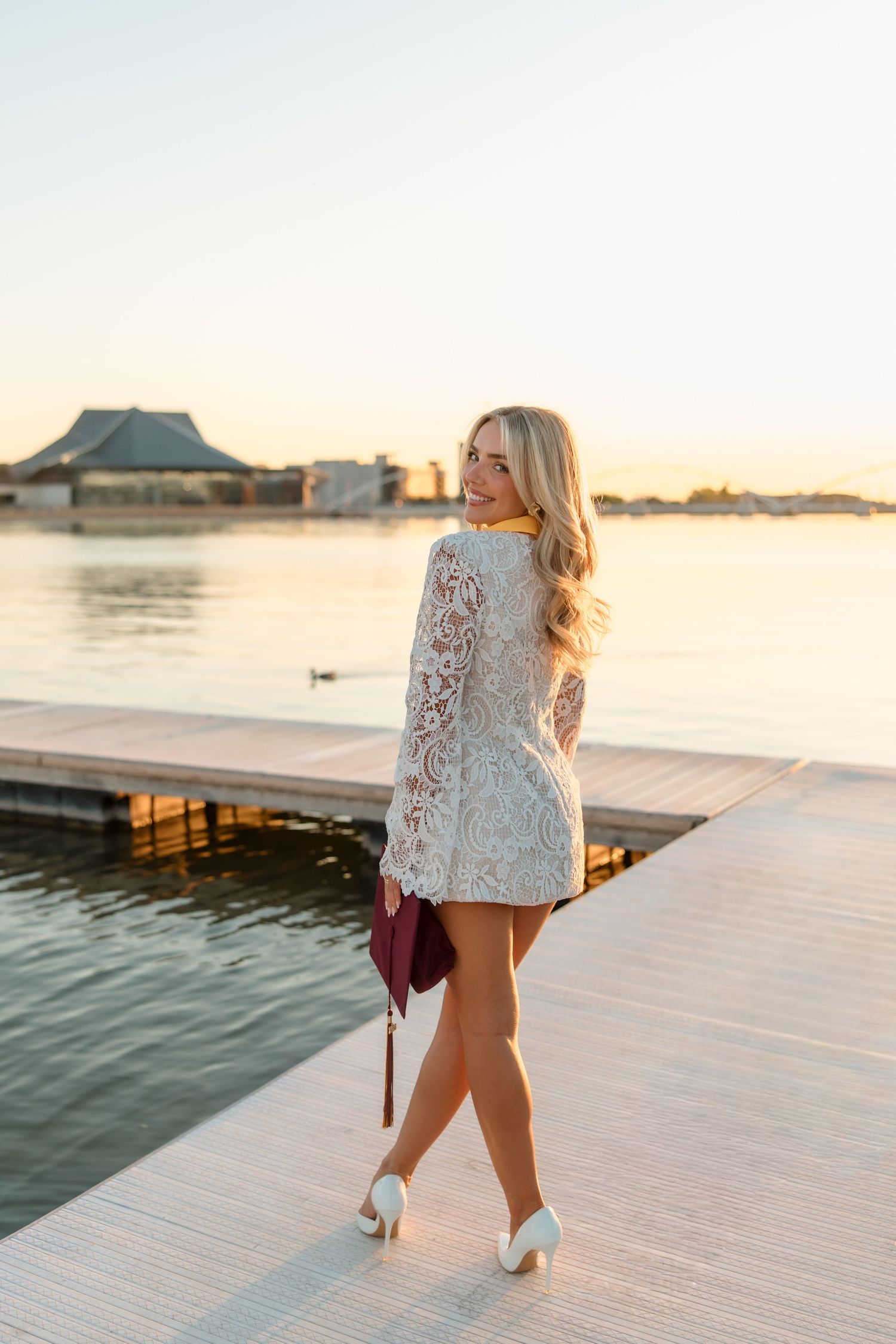 Blonde graduate in white lace dress walks along waterfront pier at sunset with diploma.