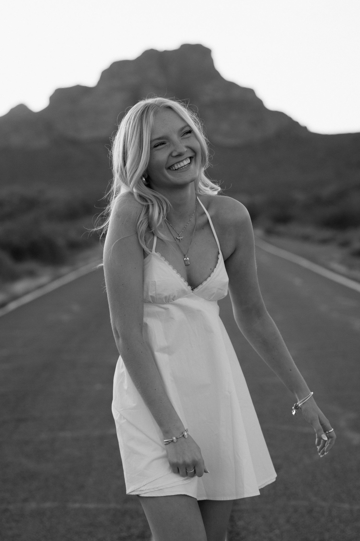 Black and white photo of a laughing blonde woman in a white dress standing on a desert road with mountains behind her.
