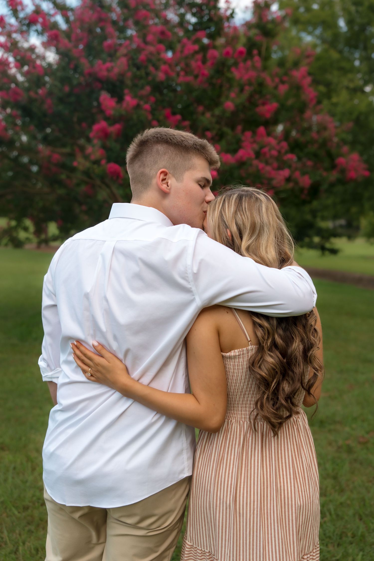 A couple shares a romantic kiss outdoors, embracing near blooming pink crepe myrtle trees on a lush green lawn.