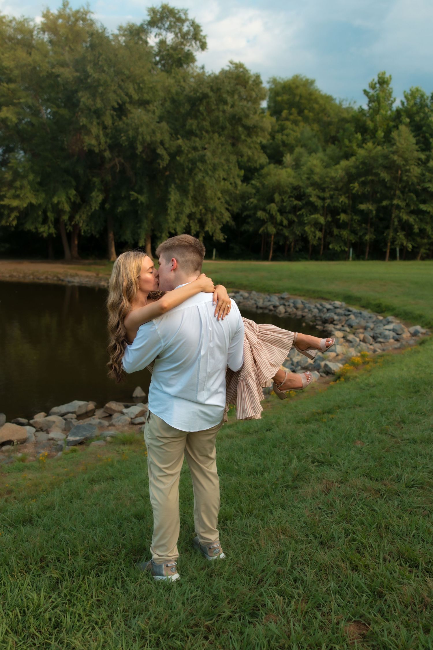 Man lifting woman in his arms near a scenic pond surrounded by lush green trees during a romantic outdoor photo session.