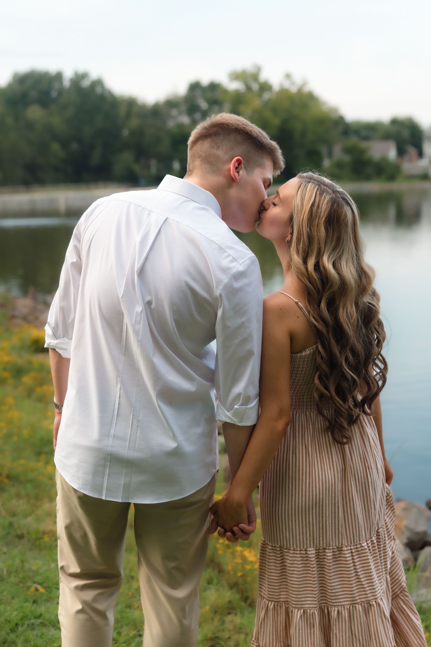 Couple kissing and holding hands by a serene lake, man in white shirt and woman in striped dress.
