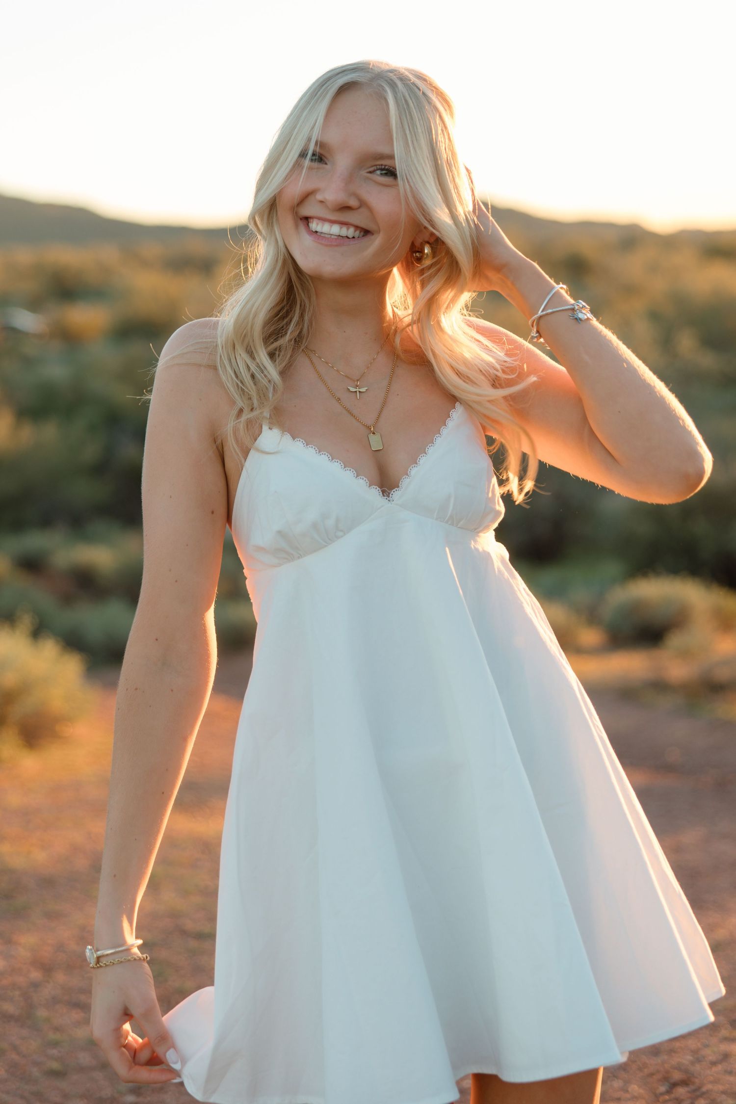 Radiant blonde woman in white mini dress posing in glowing sunset desert landscape.