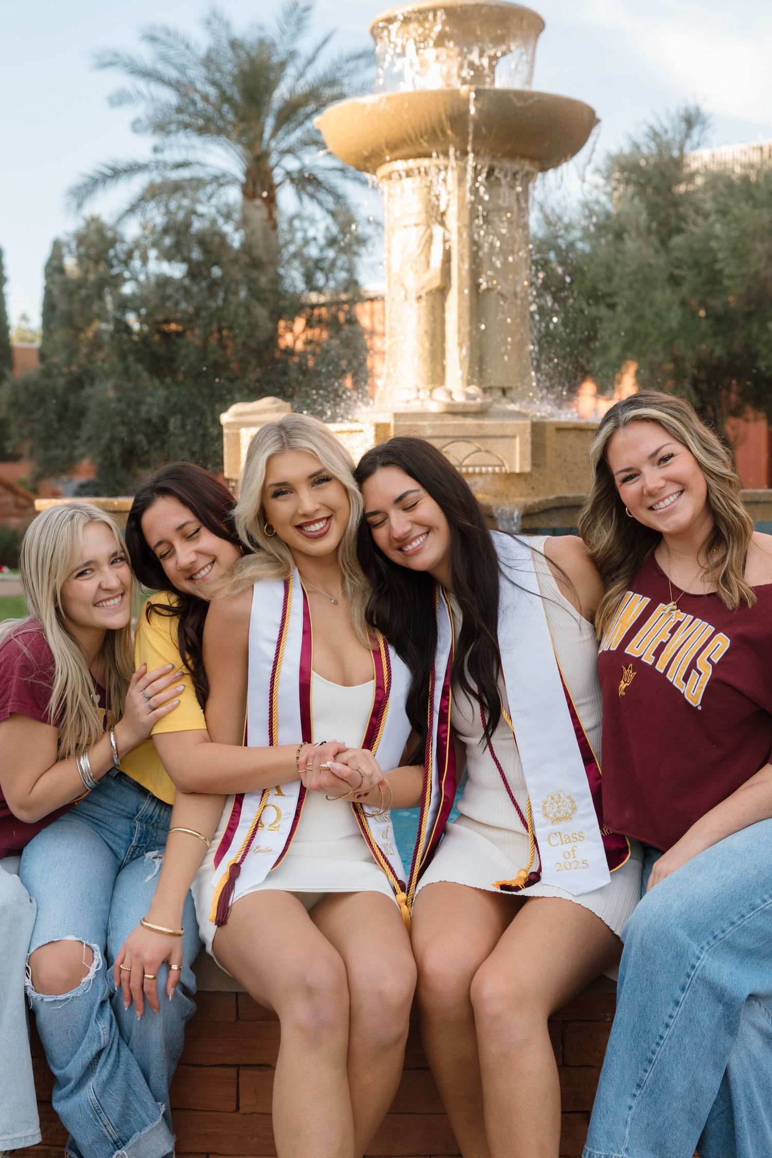 Five young women celebrating ASU graduation, sitting together laughing near a decorative fountain on campus.