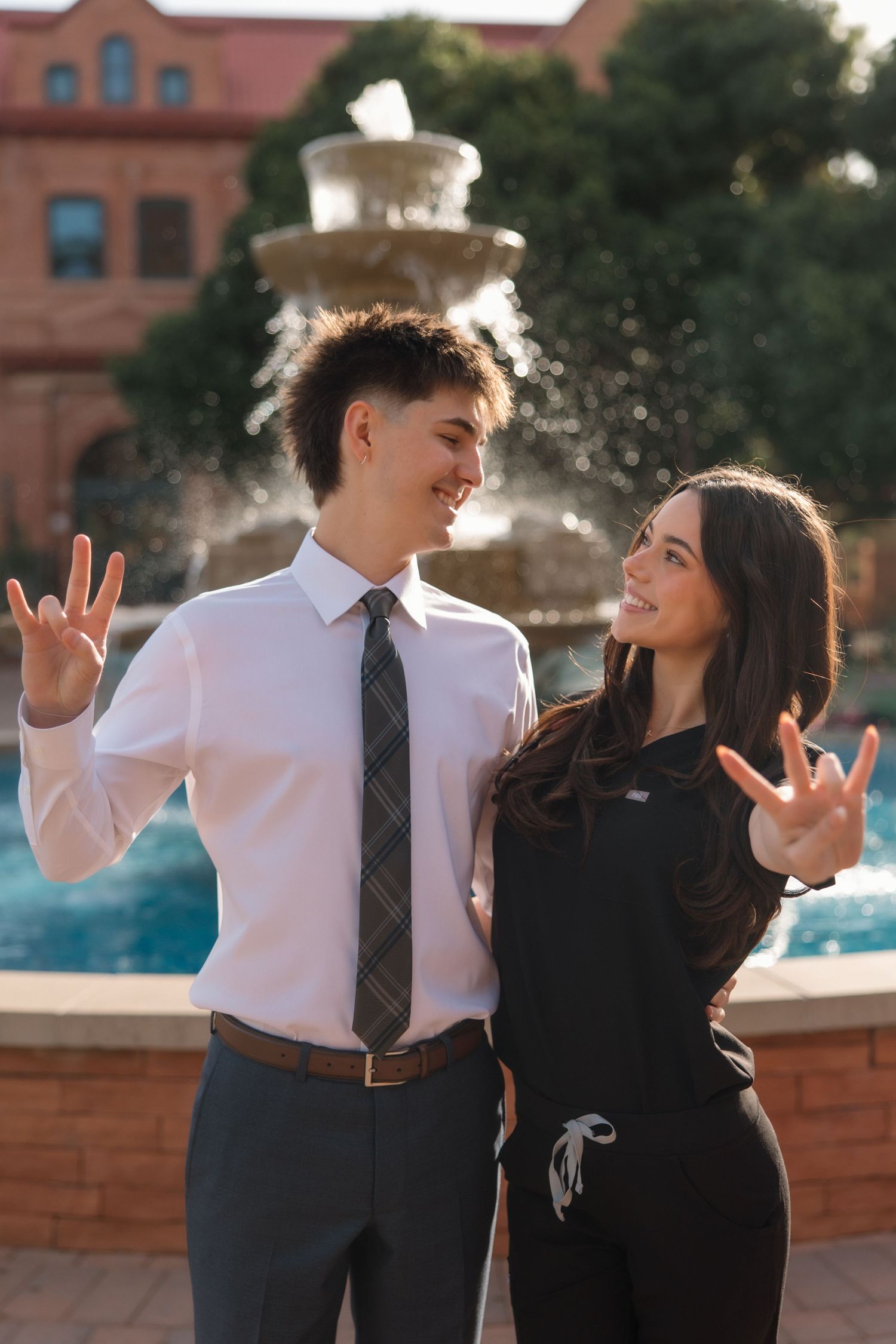 Young couple flashing hand signs in front of a fountain on a sunny university campus.