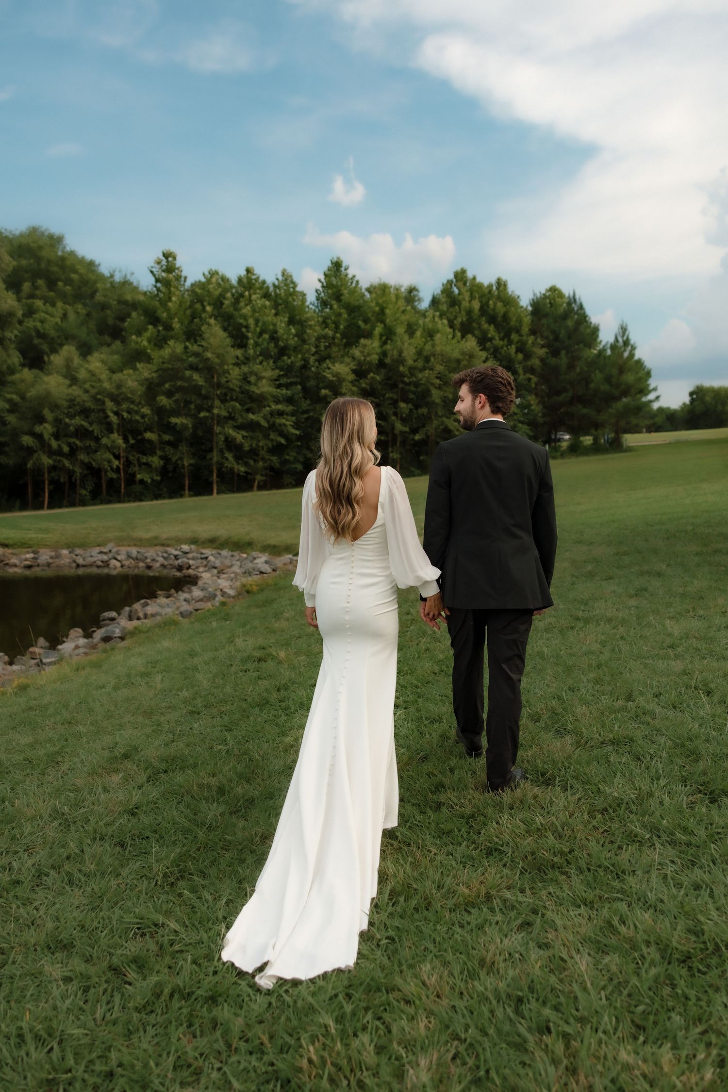 Bride and groom walking hand in hand across green lawn near a lake in elegant wedding attire.