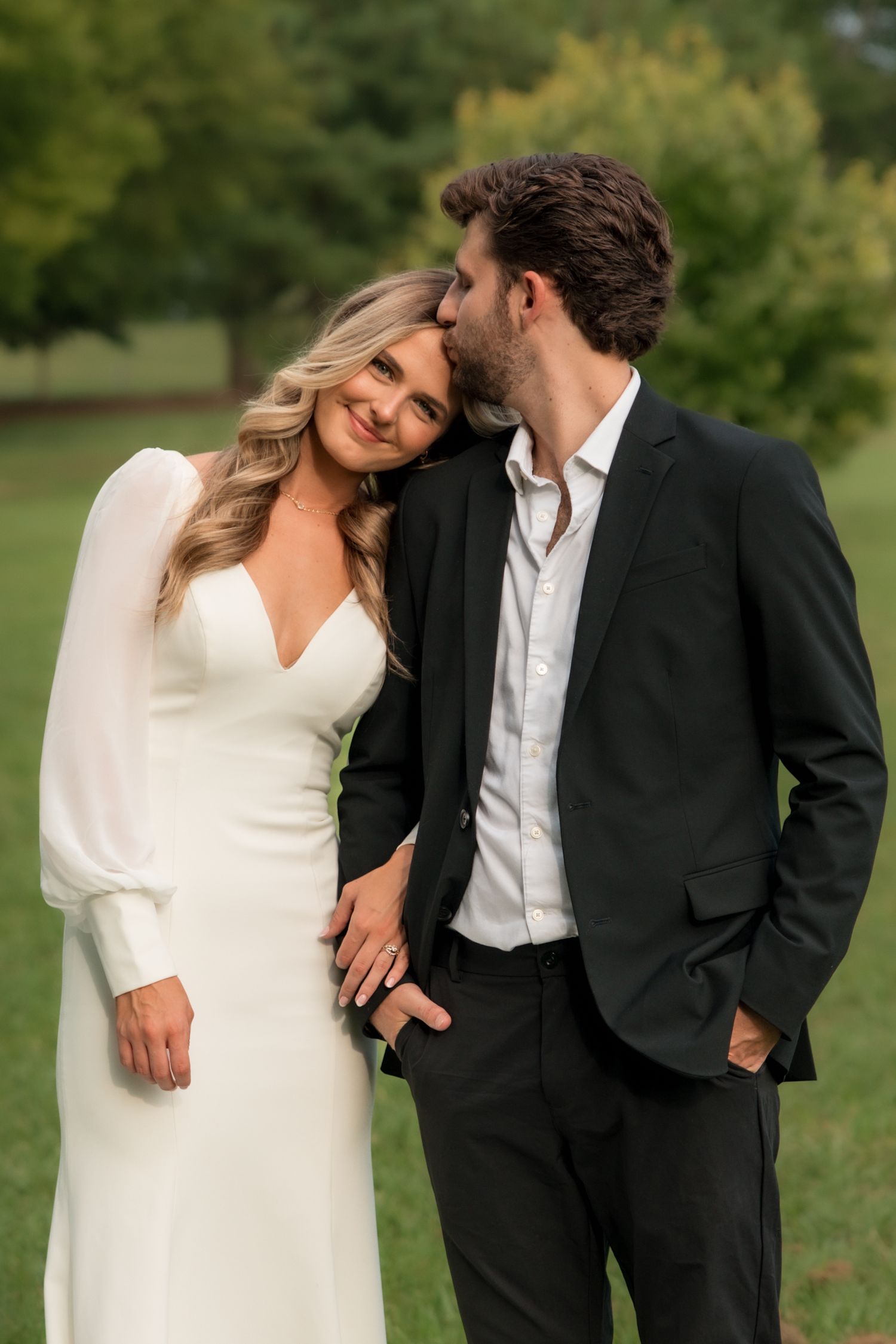 A bride in a white gown leans her head on a groom in a black suit as he kisses her forehead outdoors.
