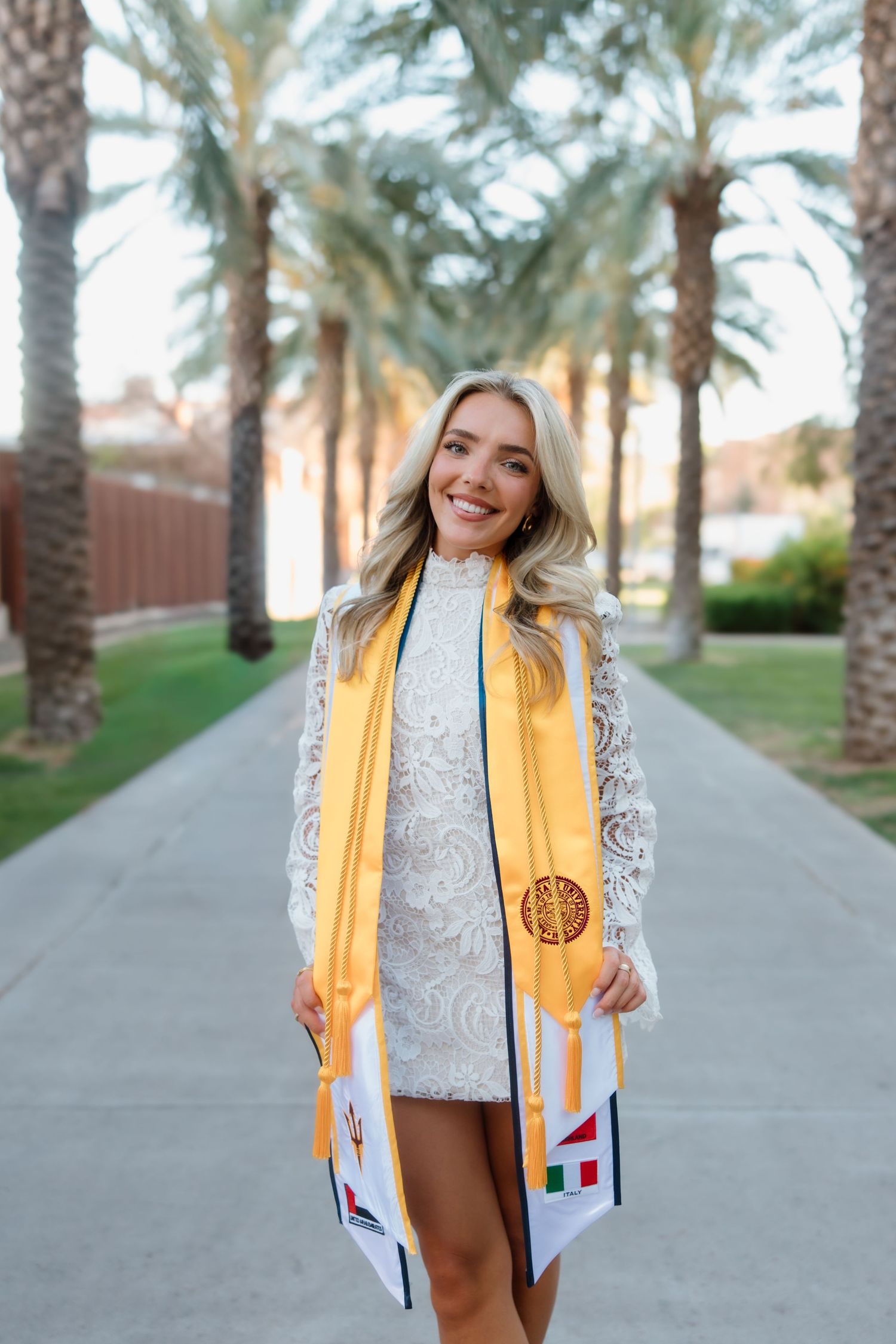 Smiling blonde graduate in white lace dress with gold ASU stole poses among palm trees.