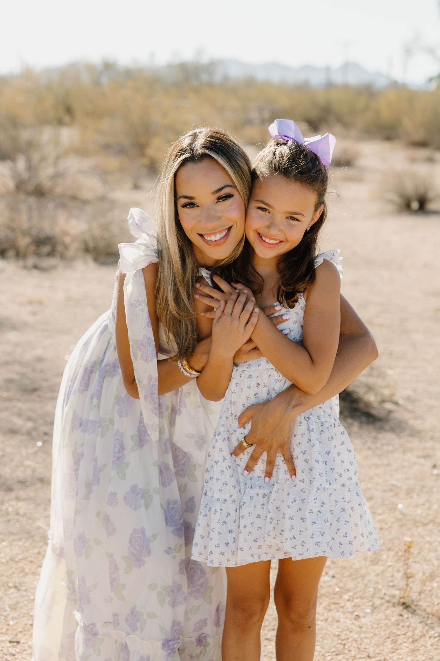 A woman and young girl in matching floral dresses smile and embrace outdoors in a desert landscape.