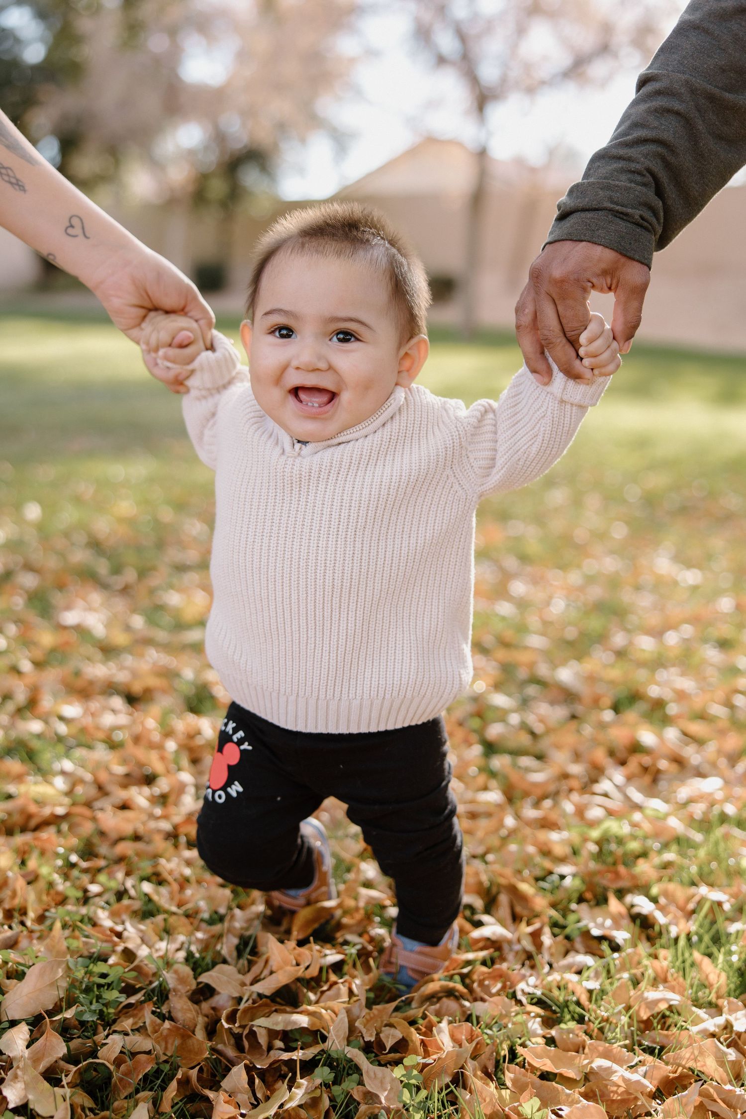 Happy baby in white sweater stands in autumn leaves, held by two adults hands in a sunny park setting.