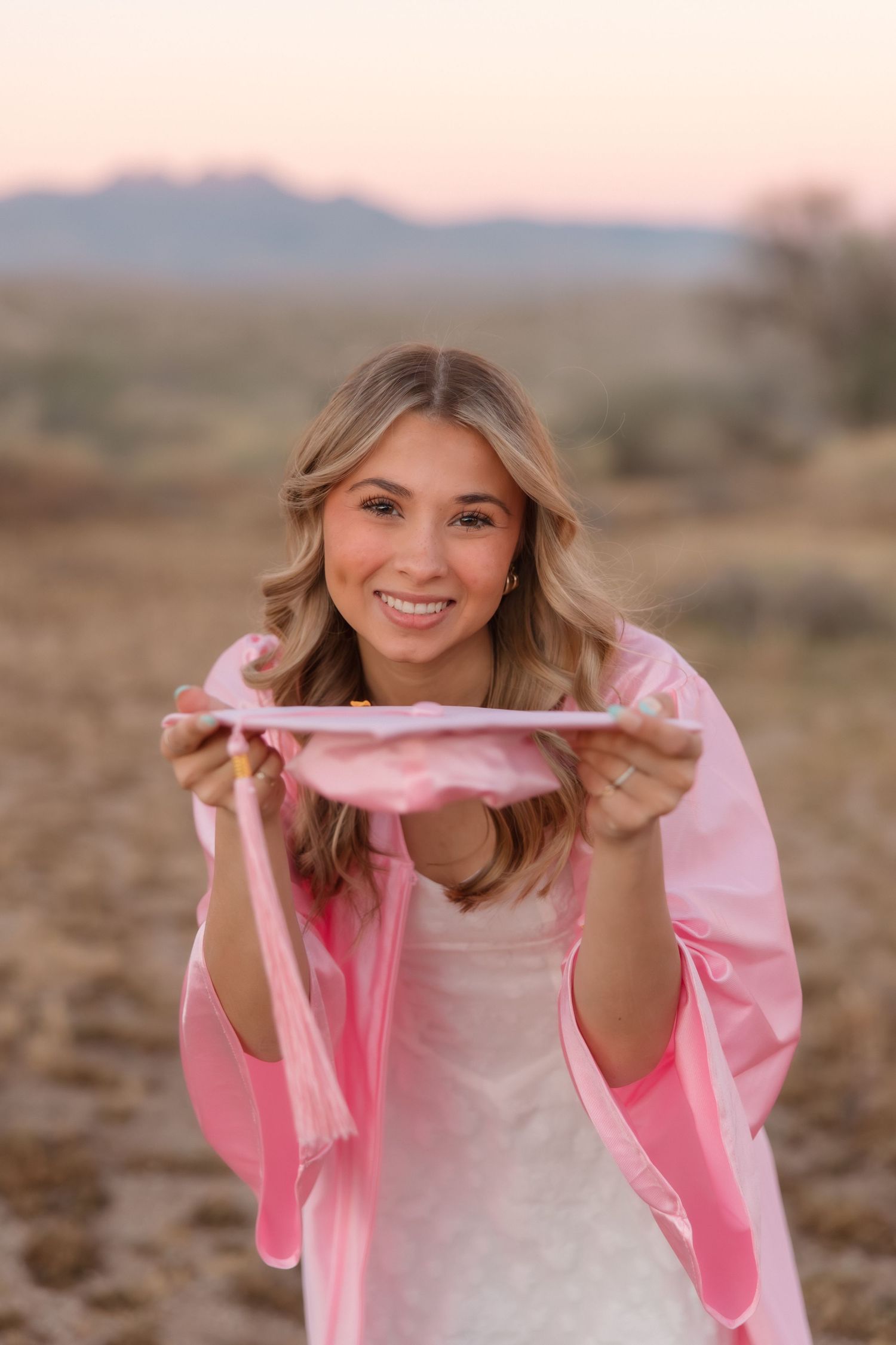 Smiling young woman in pink jacket holding pink fabric in desert landscape at sunset.