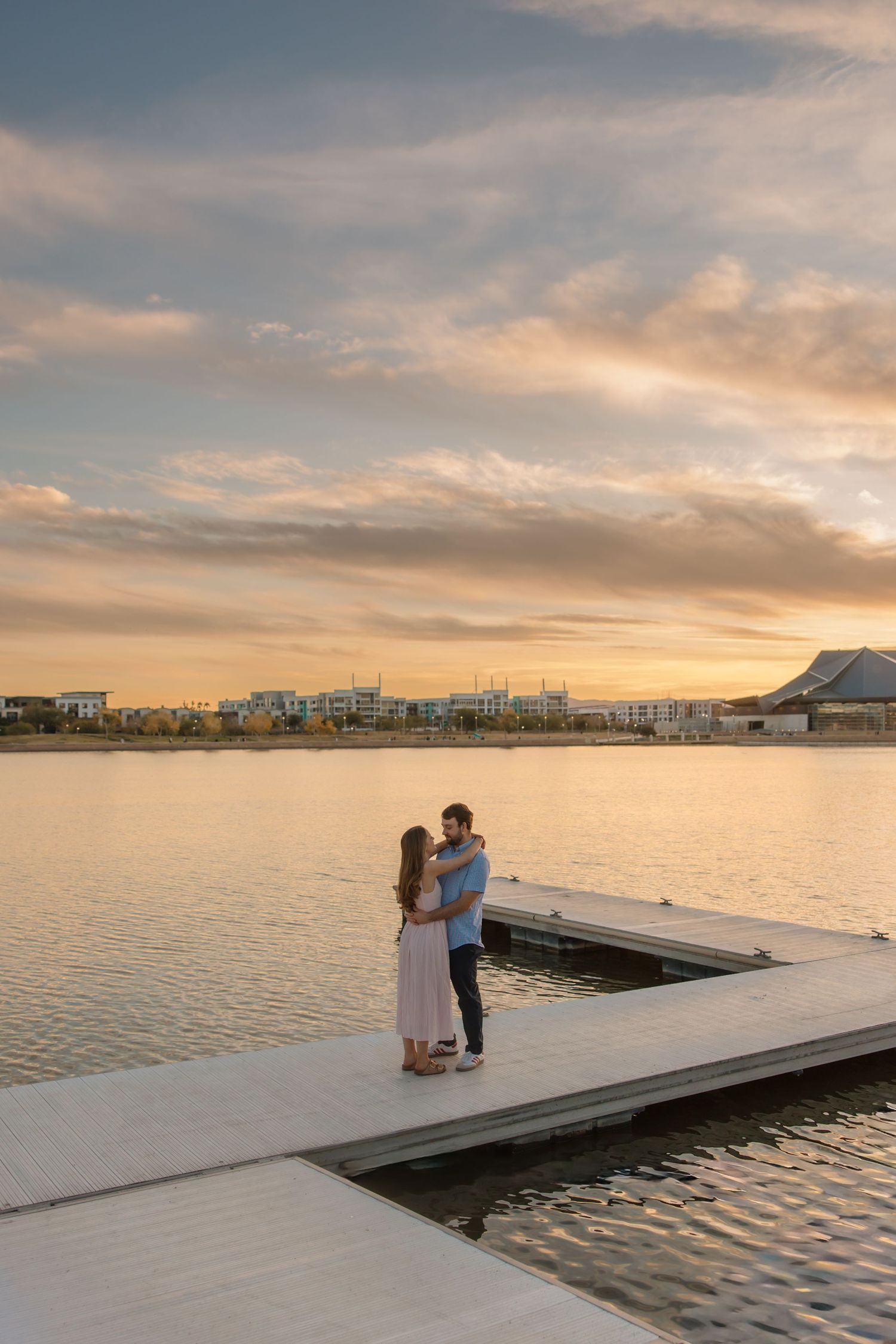 Couple embracing on dock at dramatic sunset with golden reflections on calm lake water.