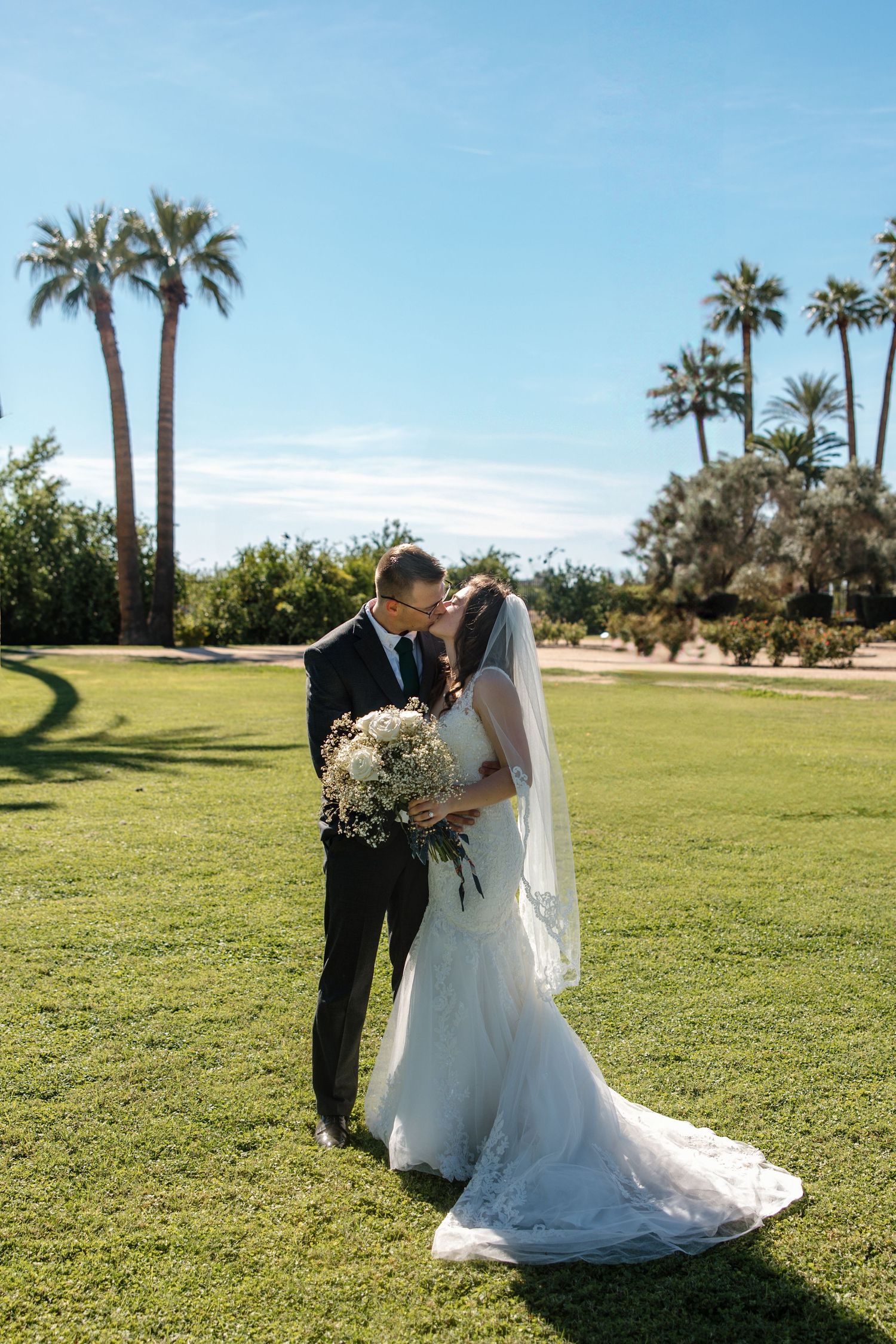 Bride and groom share a kiss on lush green lawn with tall palm trees under a bright blue sky.