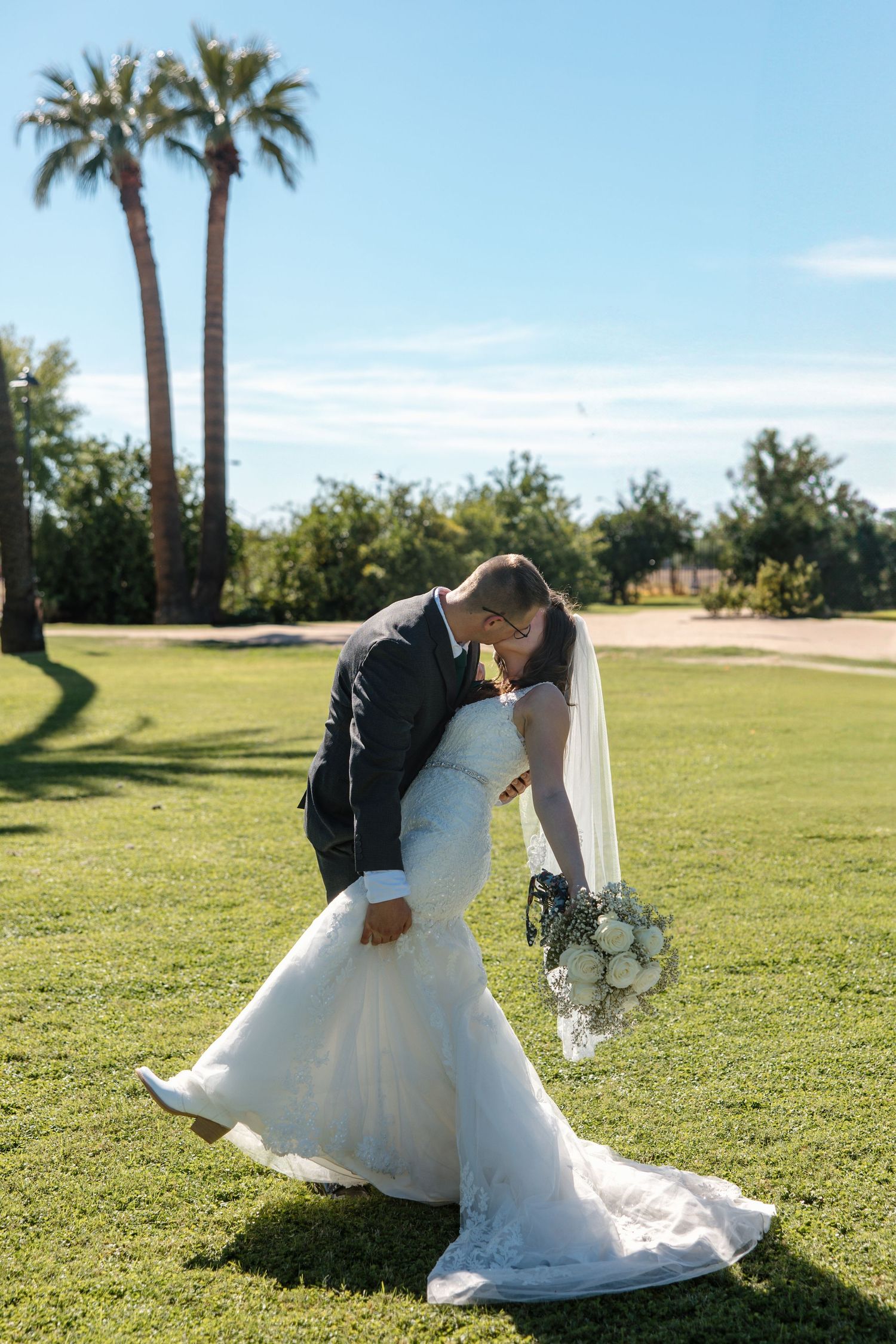 Groom dips bride for a romantic kiss on green lawn with palm trees in background.