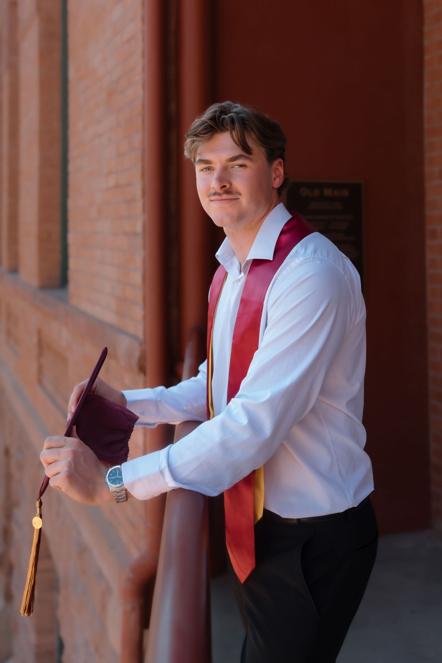 Male ASU graduate in white shirt leaning on railing with maroon cap and graduation stole.