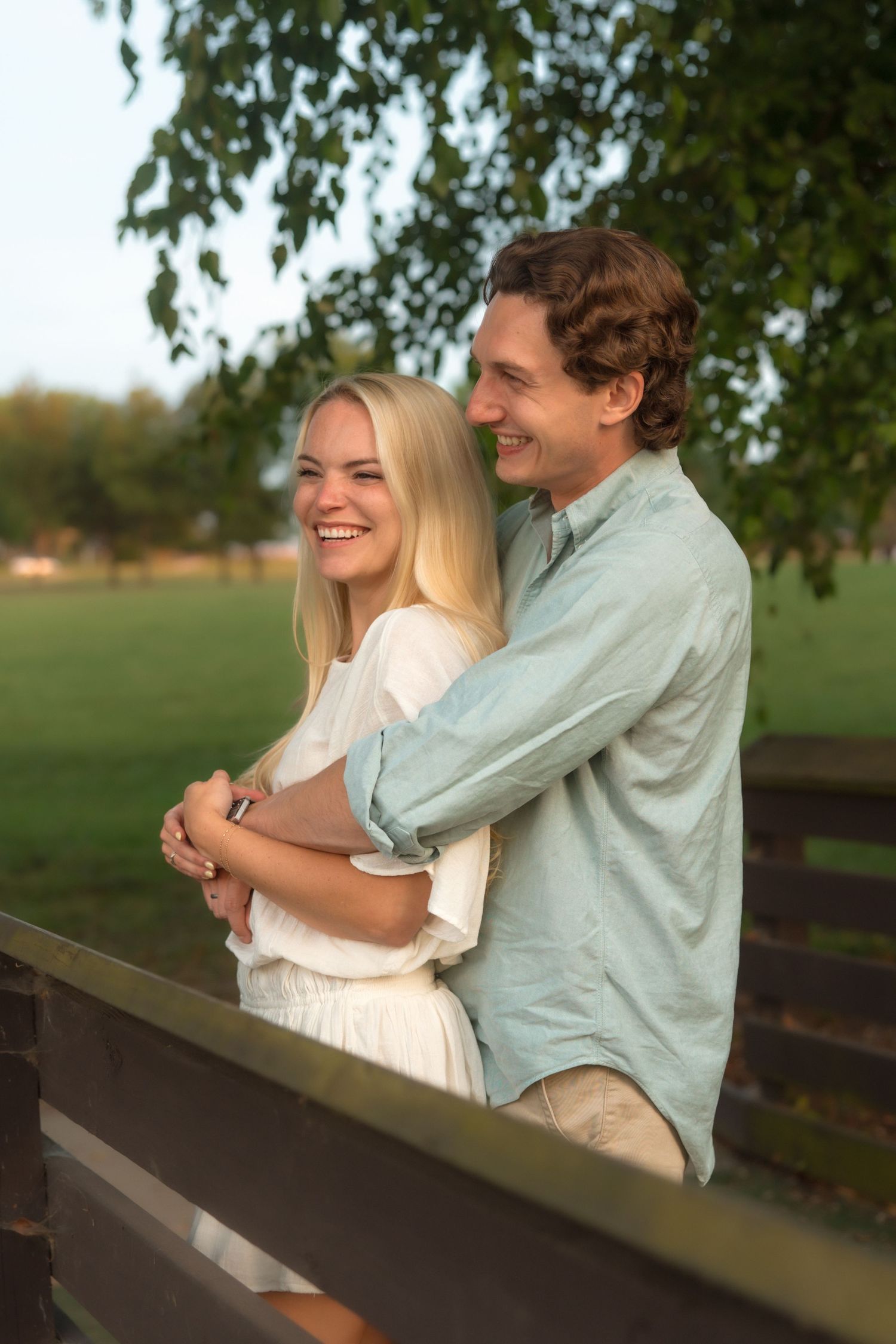 A happy couple smiles and embraces on a wooden bridge surrounded by lush green trees.