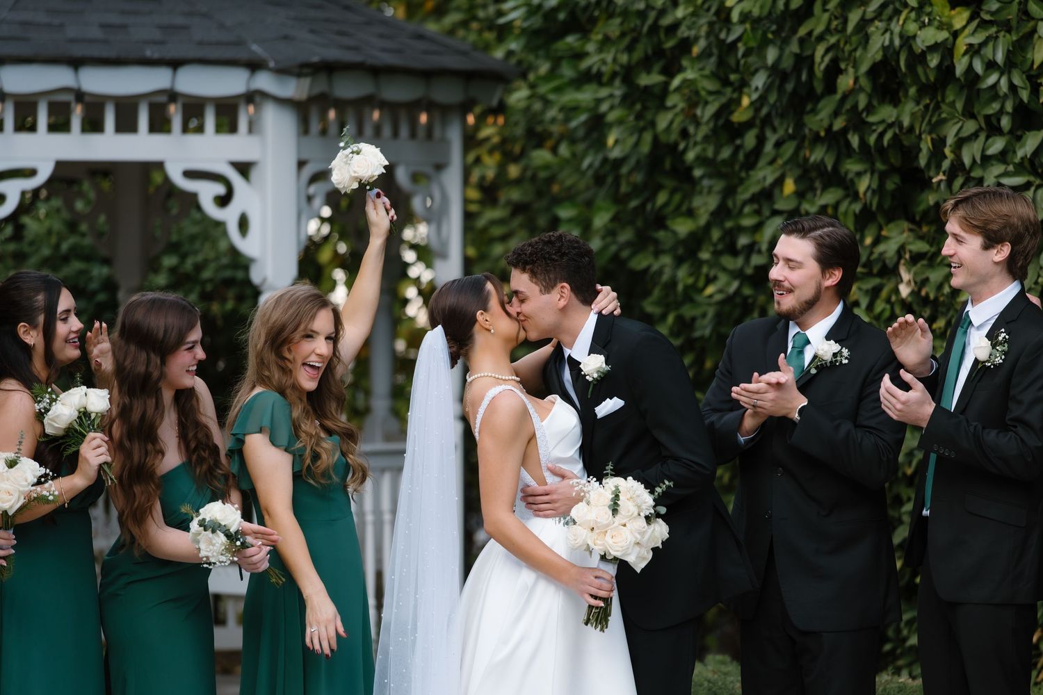 Wedding party in emerald green and black celebrates as bride and groom kiss near garden gazebo.