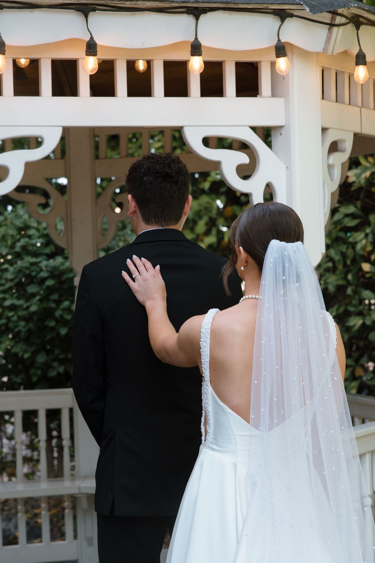 Bride places hand on groom's back as they face a white garden gazebo decorated with string lights.