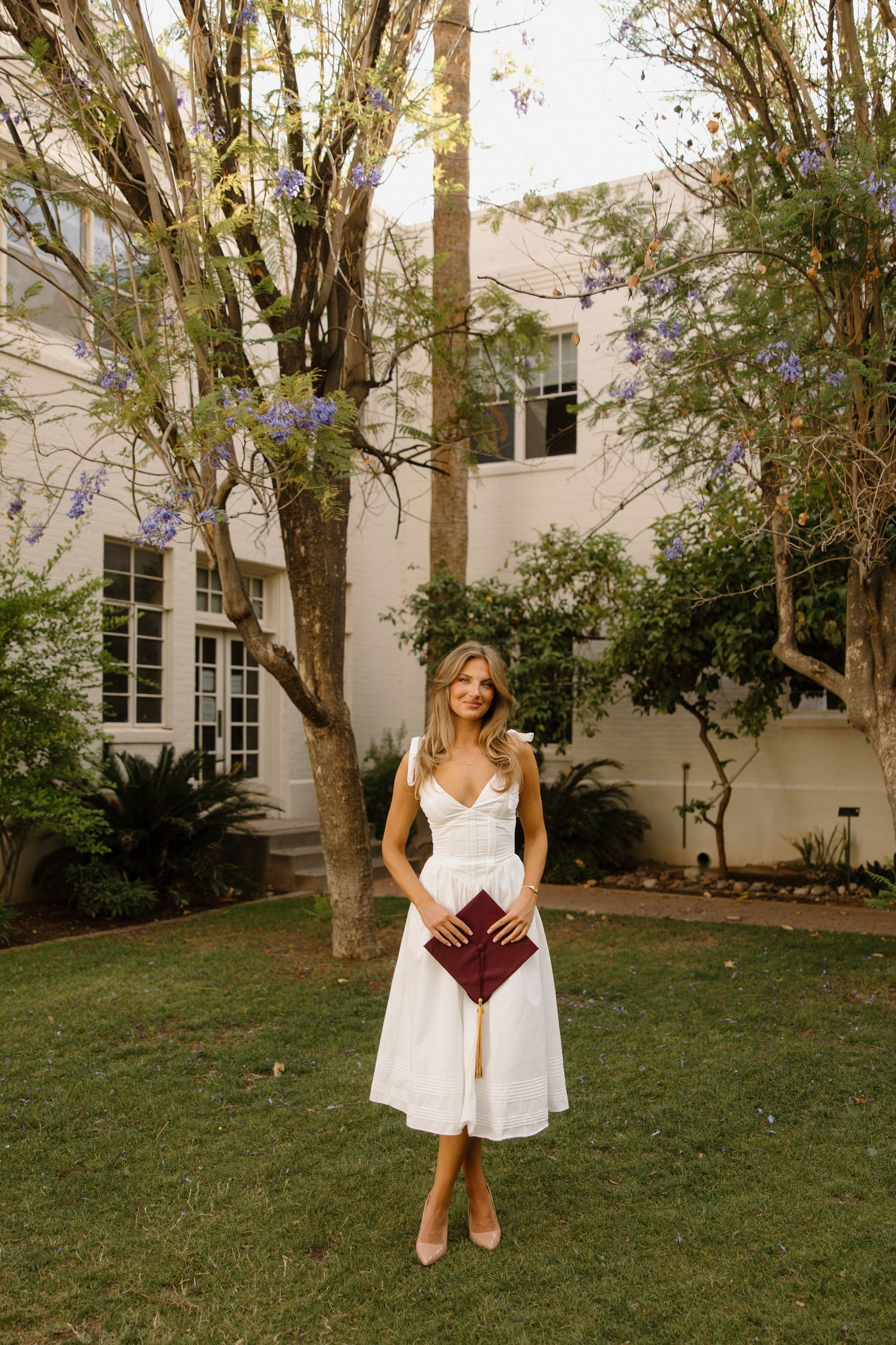 Woman in white midi dress holding burgundy clutch stands on green lawn before white building with flowering trees.