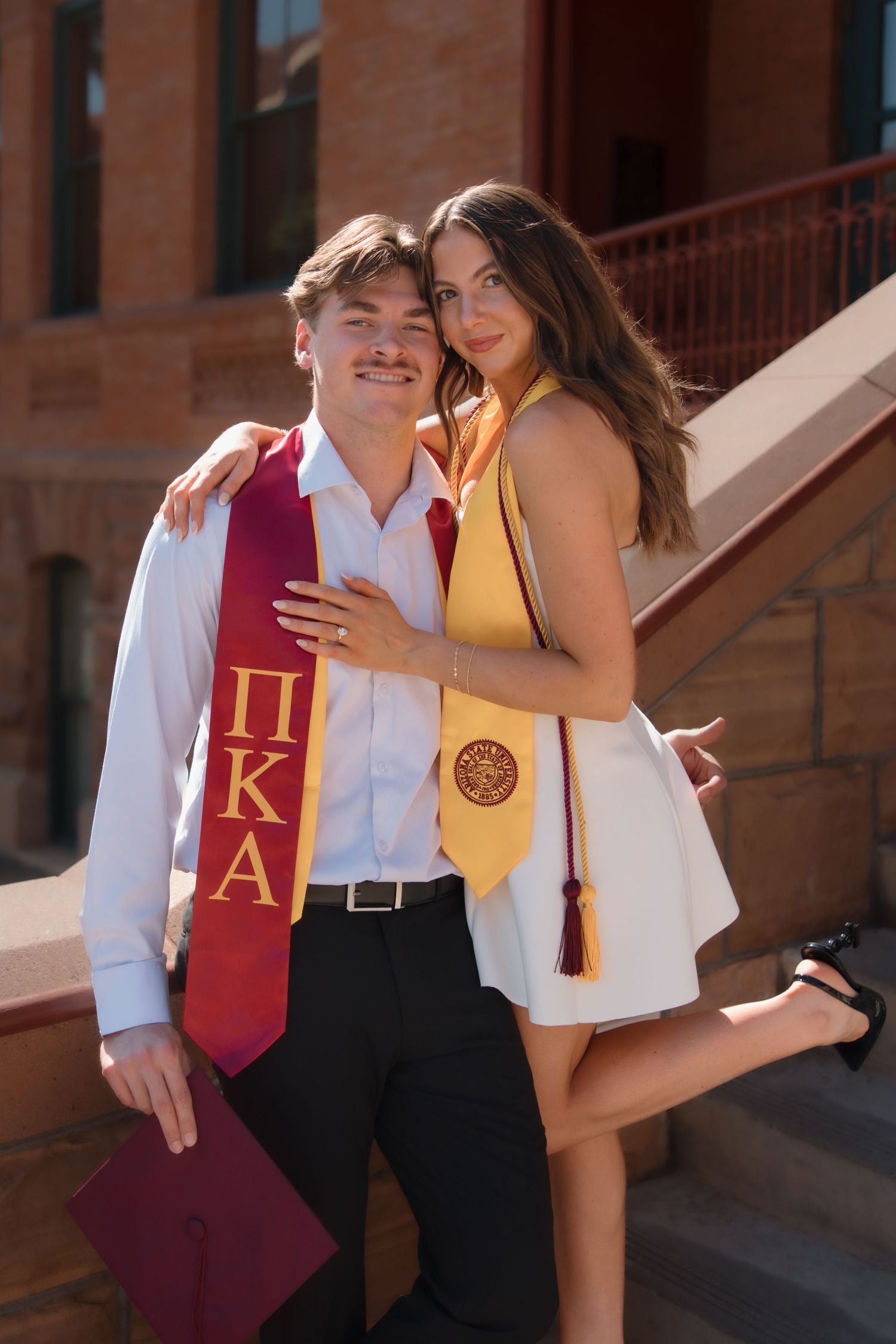 Two college graduates pose on steps wearing Greek organization stoles, smiling together in graduation attire.