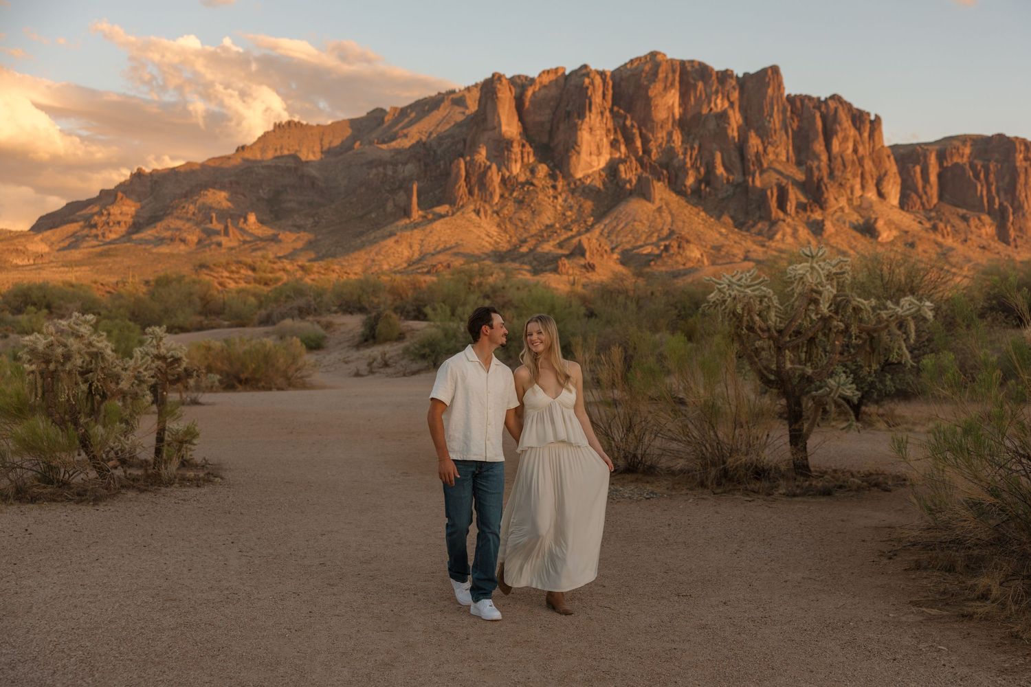 Couple walks hand-in-hand on desert path with Superstition Mountains glowing at golden hour in Arizona.