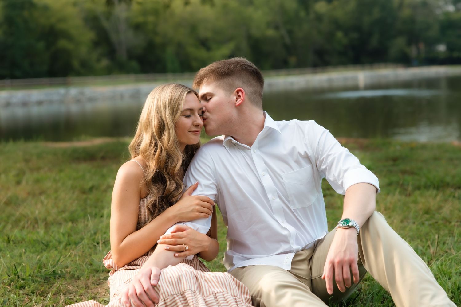 Romantic couple sitting by a serene lake, sharing a tender kiss outdoors on a sunny day.