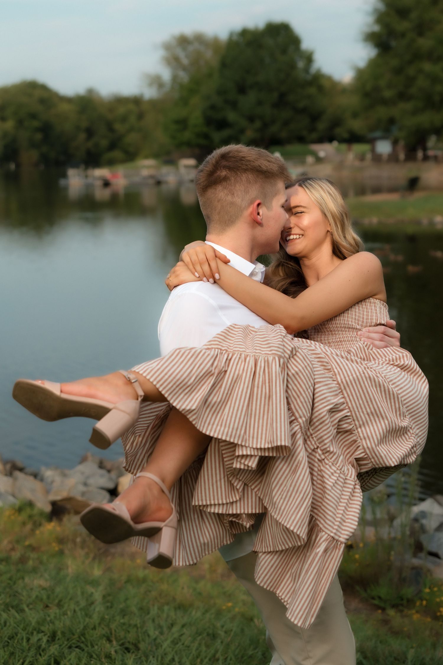 A man lifts a smiling woman in a striped dress near a serene lake, sharing a romantic moment outdoors.