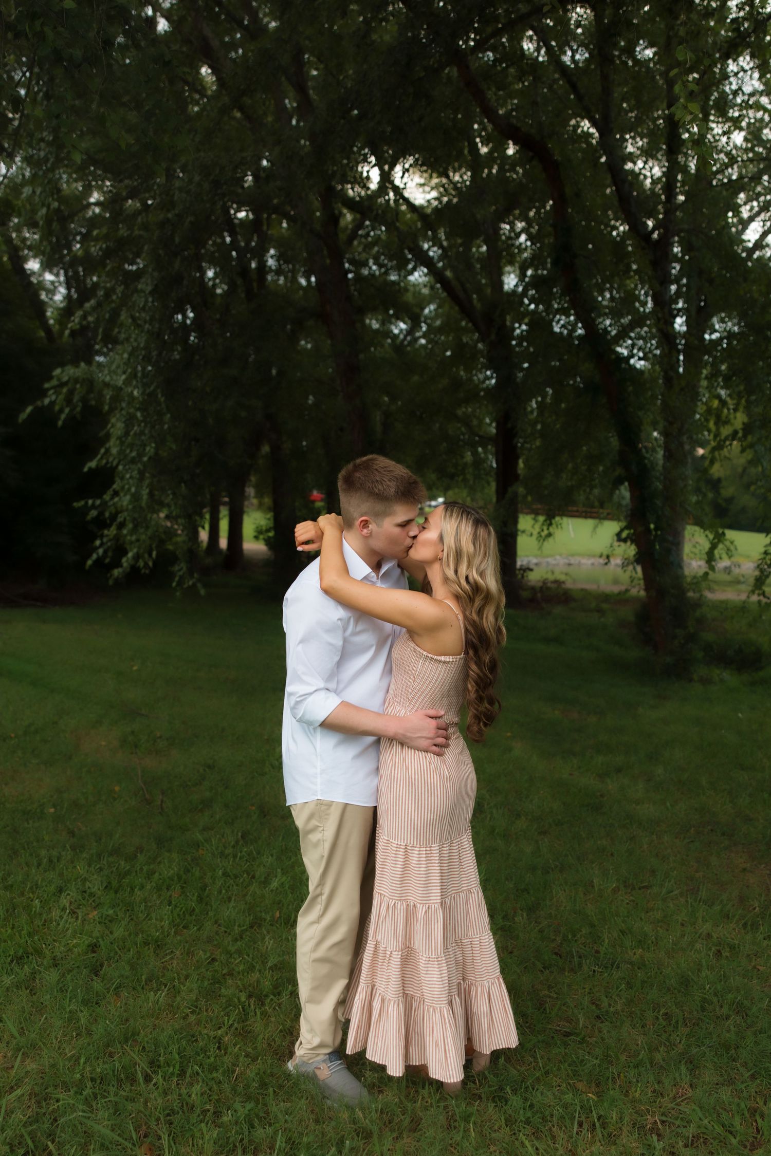 Couple sharing a romantic kiss outdoors on green grass surrounded by lush trees during golden hour.