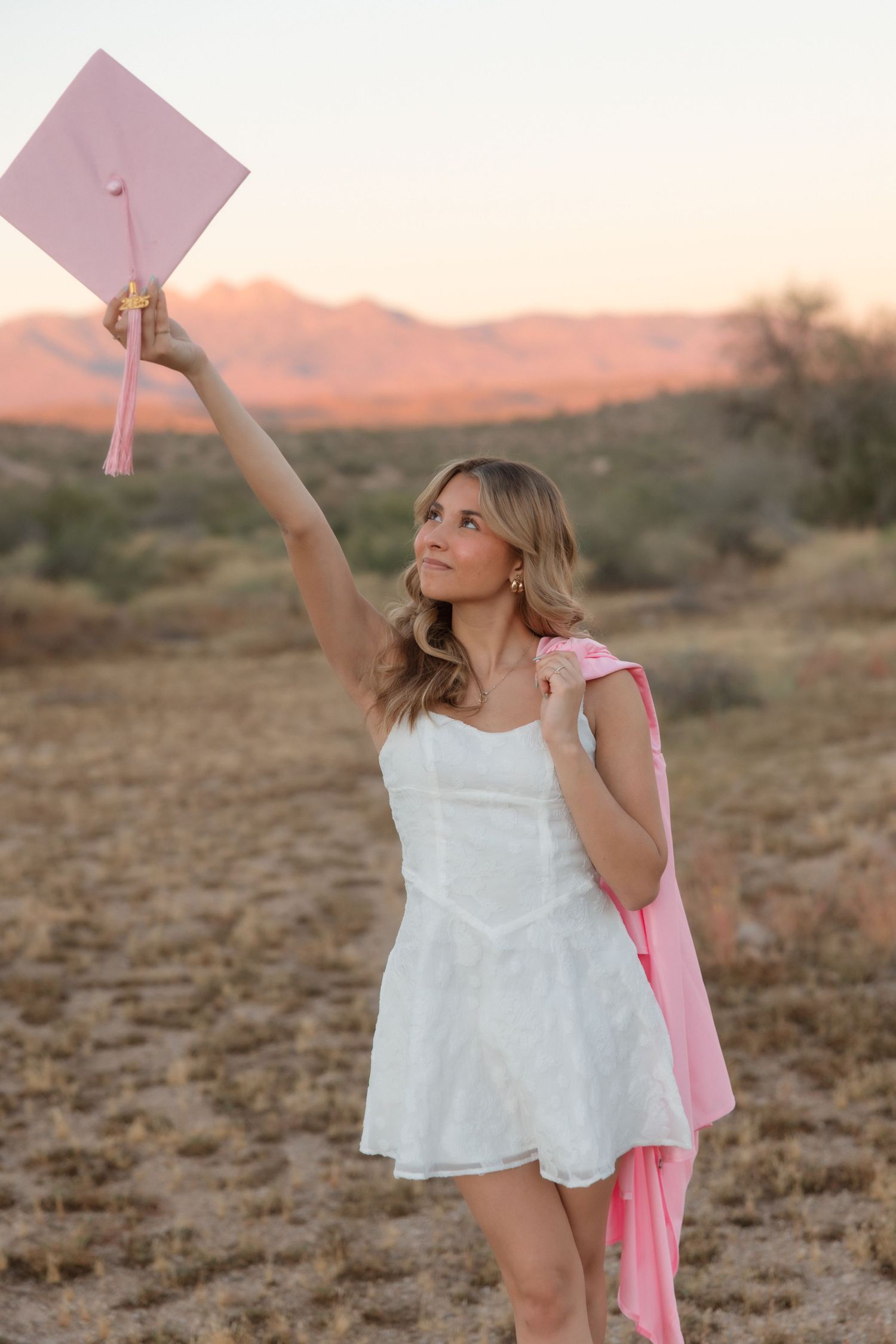 Young woman in white dress holding pink graduation cap aloft in desert landscape at sunset.