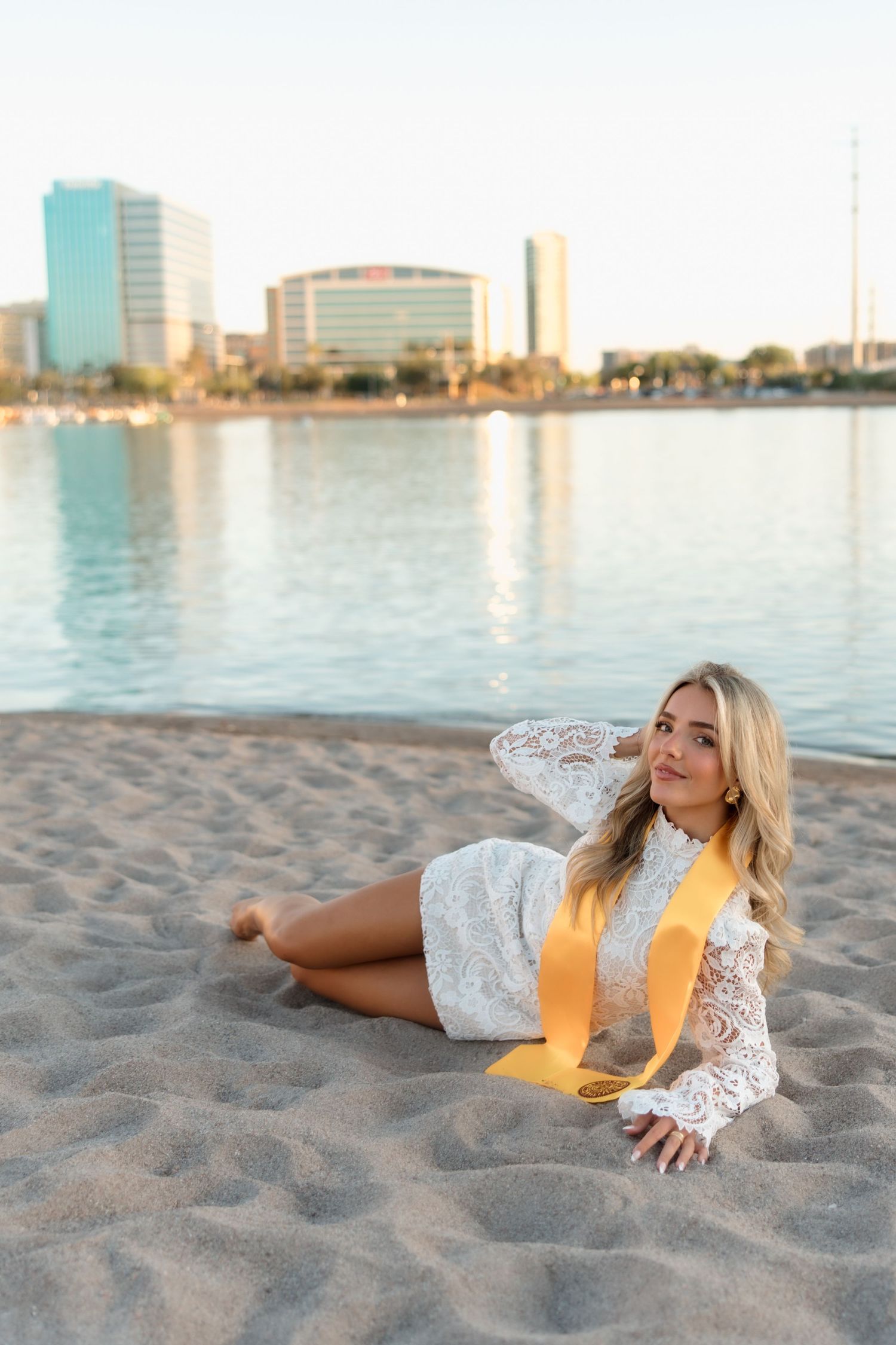 Smiling blonde graduate in white lace dress with gold stole sits on sandy urban beach.