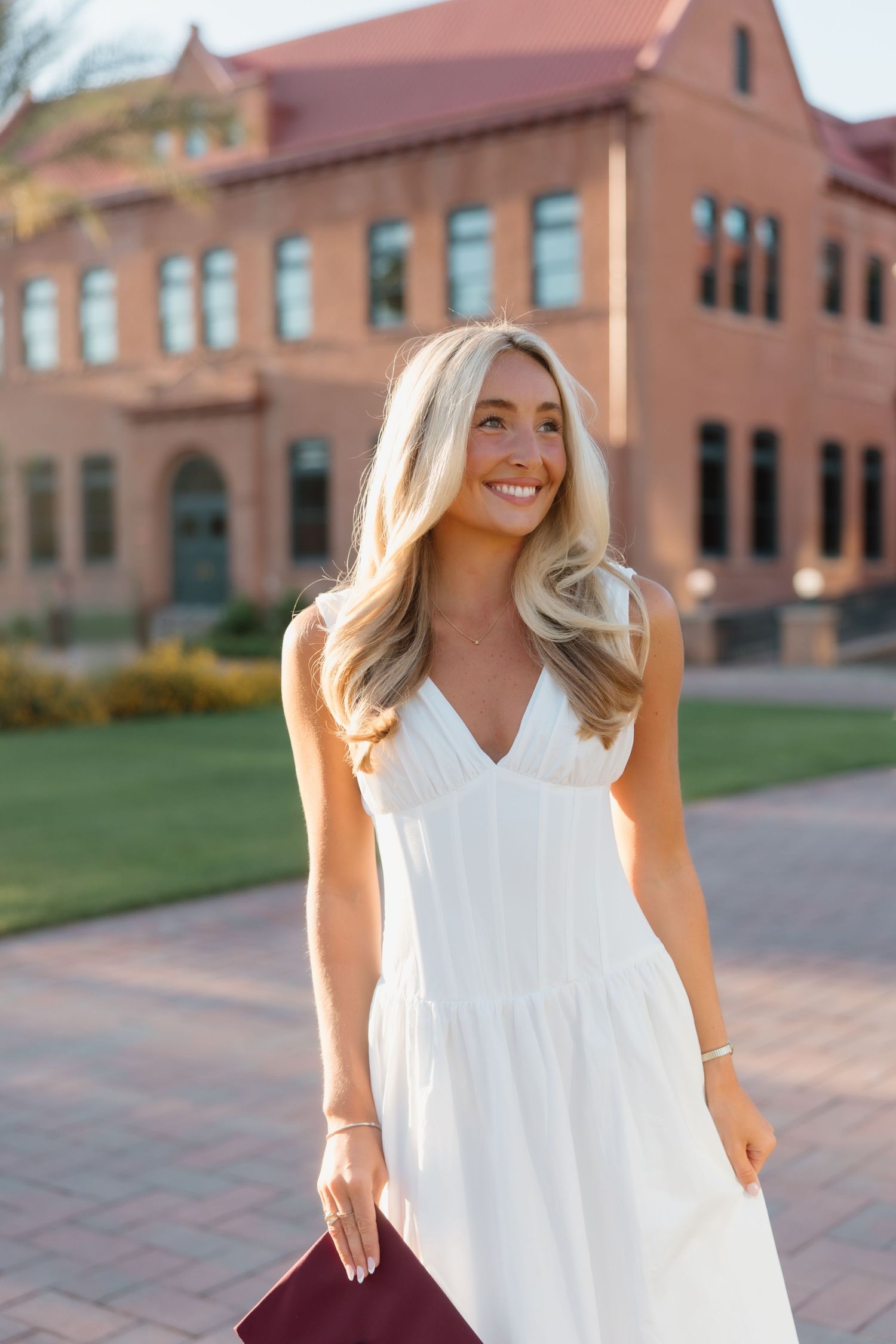 Young woman in white sundress holding maroon folder, smiling outdoors near brick university building.