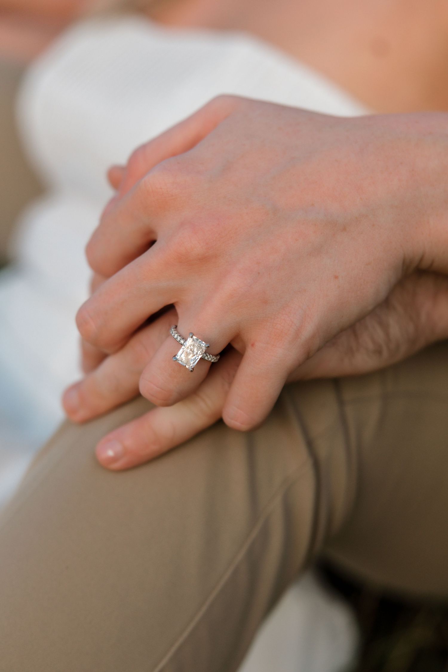 Close-up of a large radiant-cut diamond engagement ring on a womans hand resting on her partners knee.