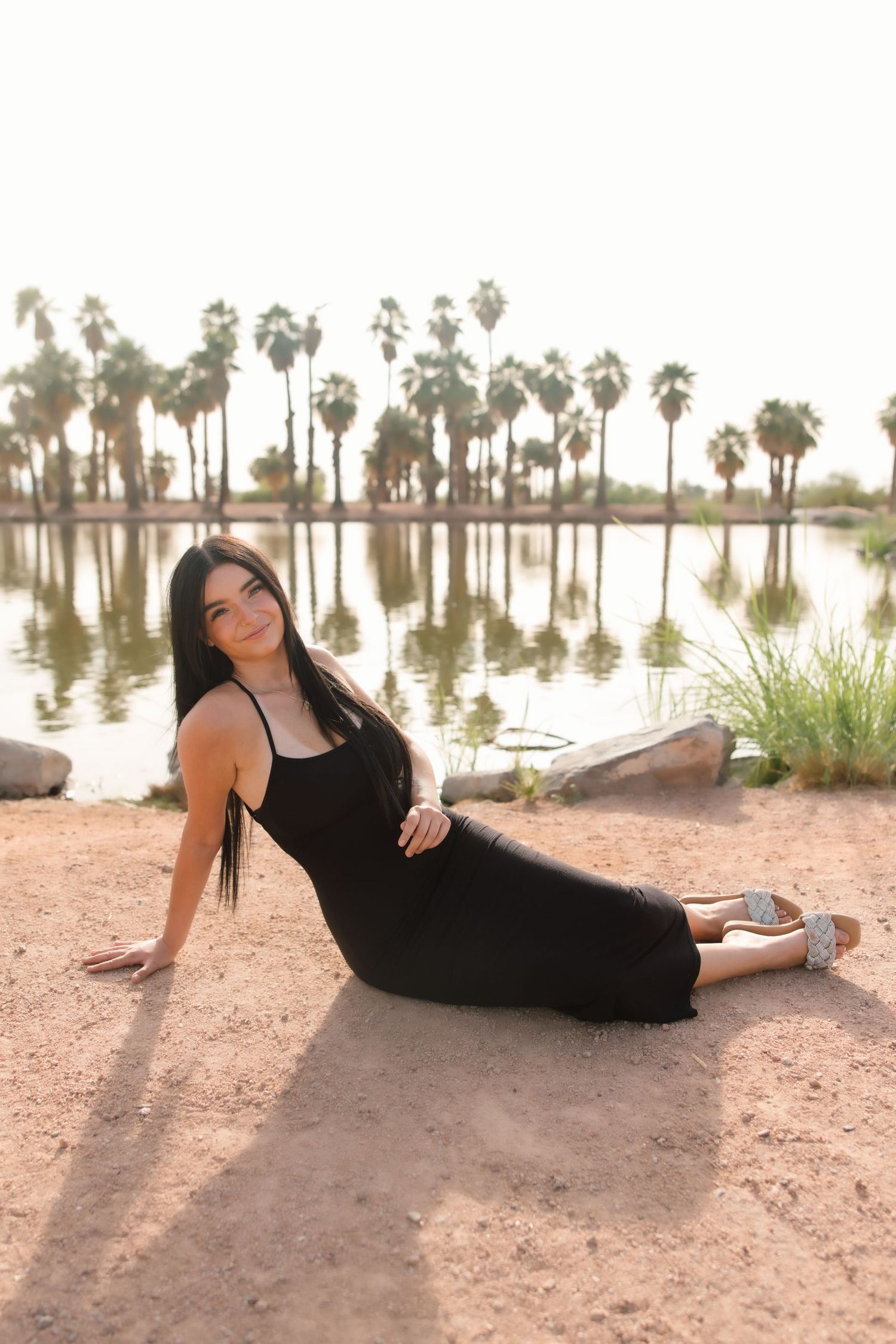 Dark-haired woman in black dress reclines gracefully by palm tree-lined lake at sunset.