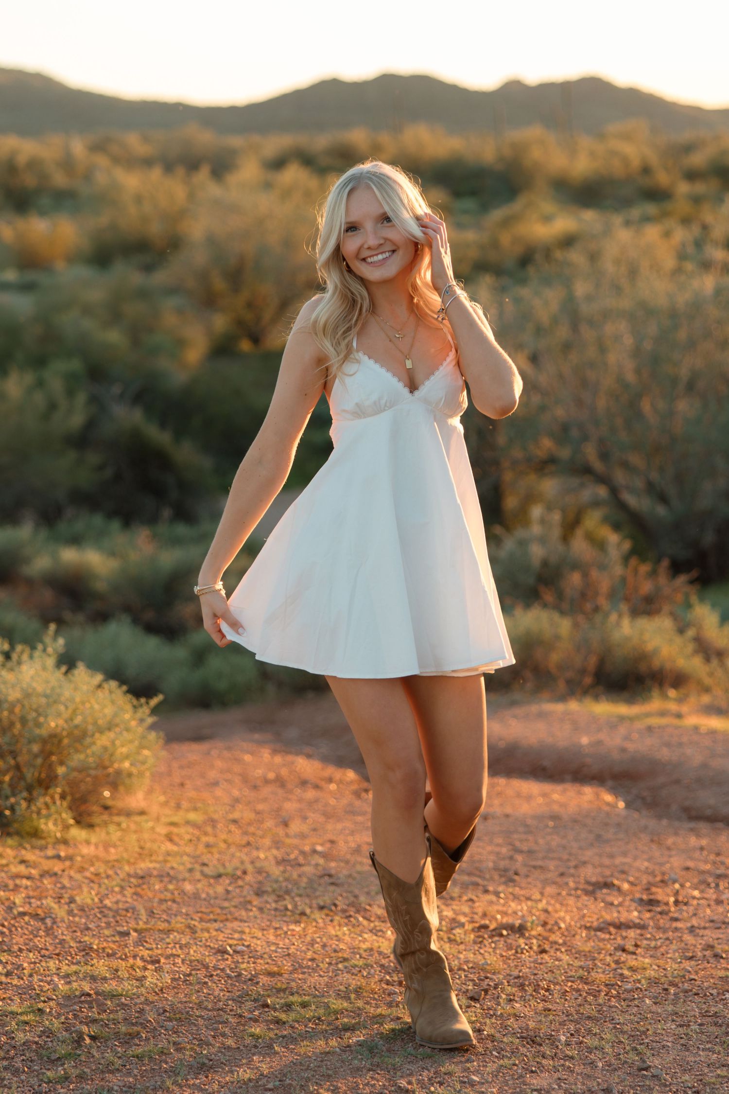 Smiling blonde woman in white mini dress and cowboy boots posing in Arizona desert at golden sunset.