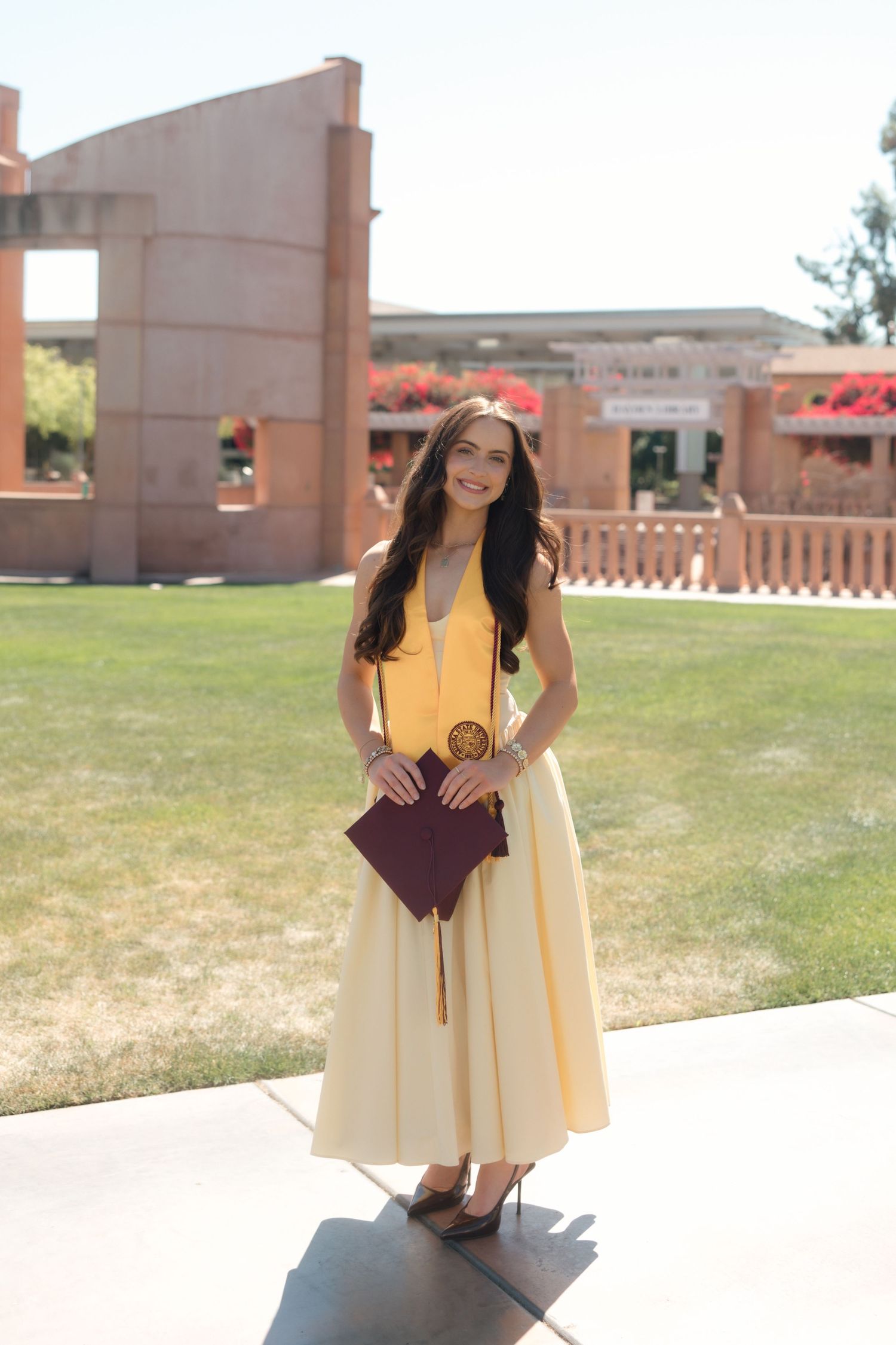 Young woman in yellow dress holding maroon graduation cap smiles outdoors on sunny college campus.