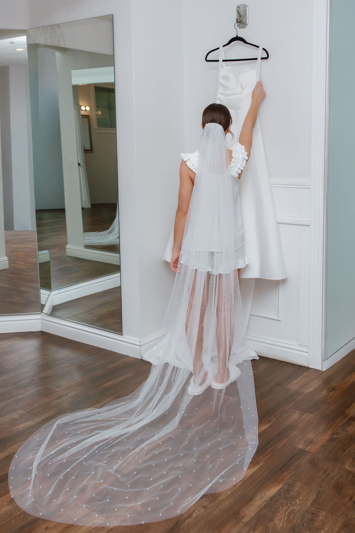 Bride in white veil reaching up to touch her wedding dress hanging on a wall next to a large mirror.