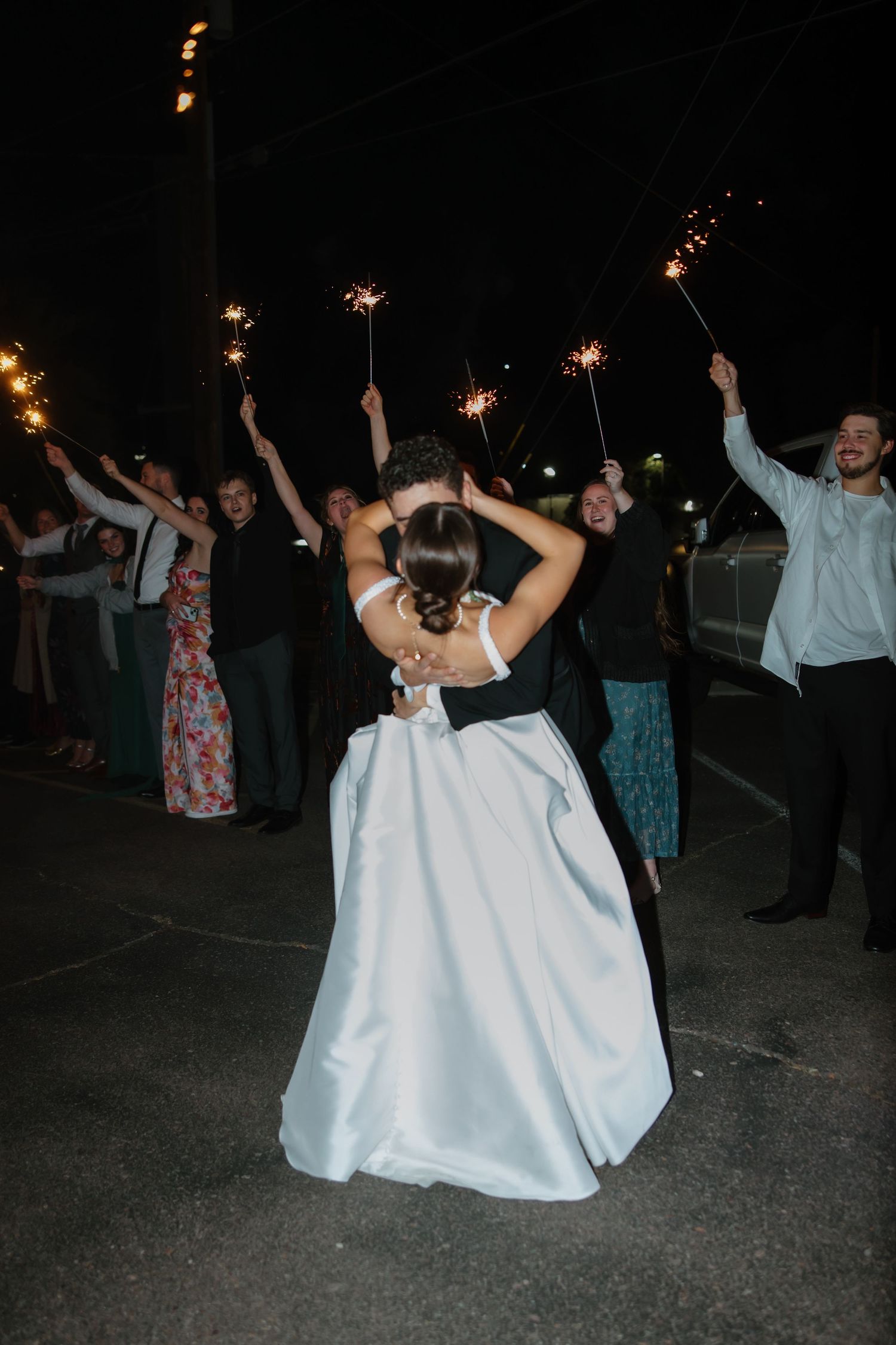 Couple shares tender kiss during sparkler exit surrounded by celebrating wedding guests holding lit sparklers at night.