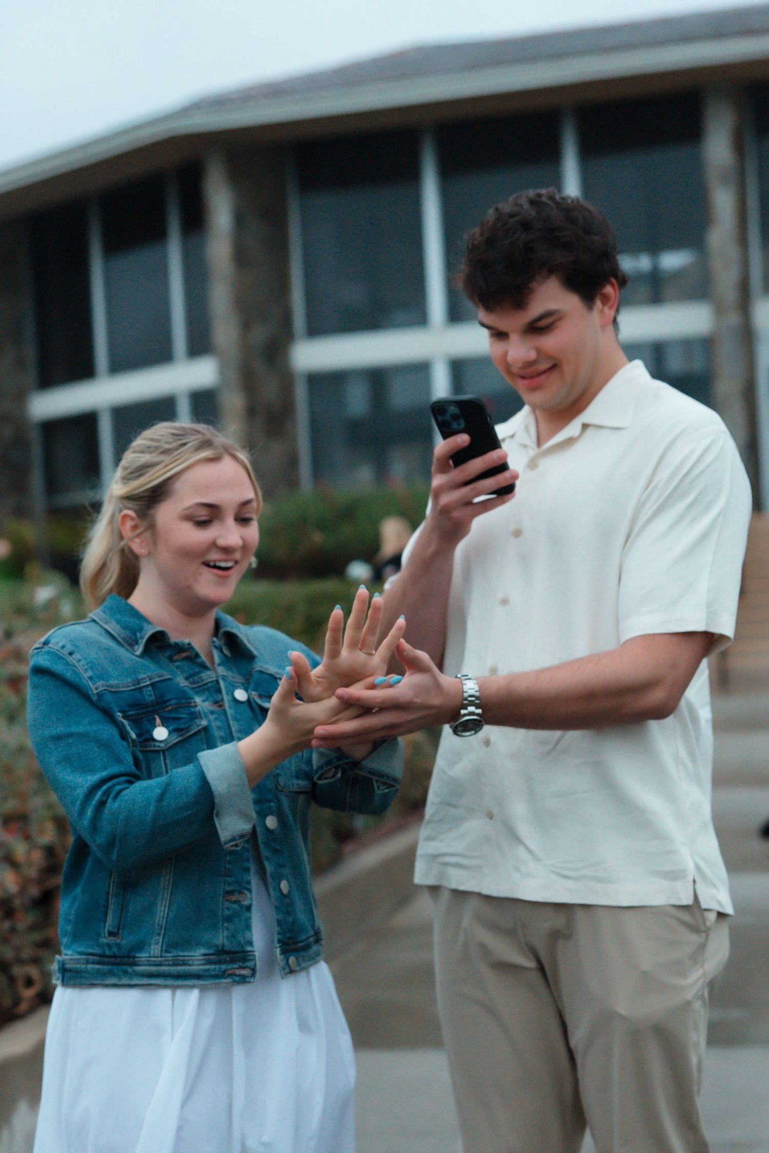 Newly engaged couple admiring the engagement ring while taking a photo on a smartphone outdoors.