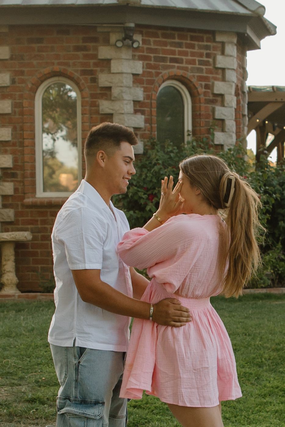 A young couple embraces and smiles at each other outdoors near a brick building, woman in pink dress, man in white shirt.