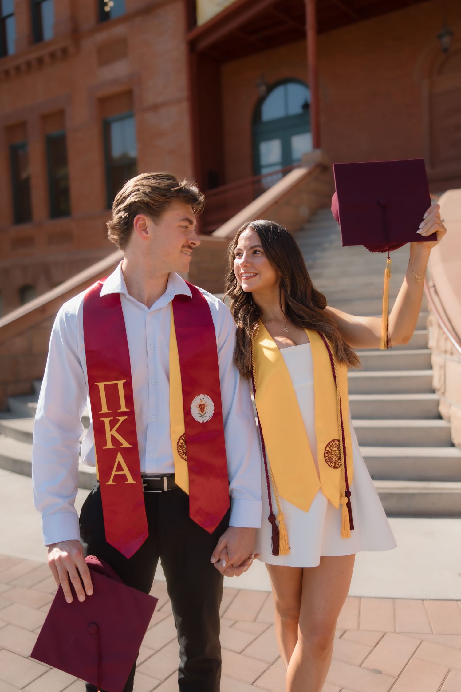 Happy graduating couple holding hands wearing Pi Kappa Alpha stole and gold honor stole outside university.