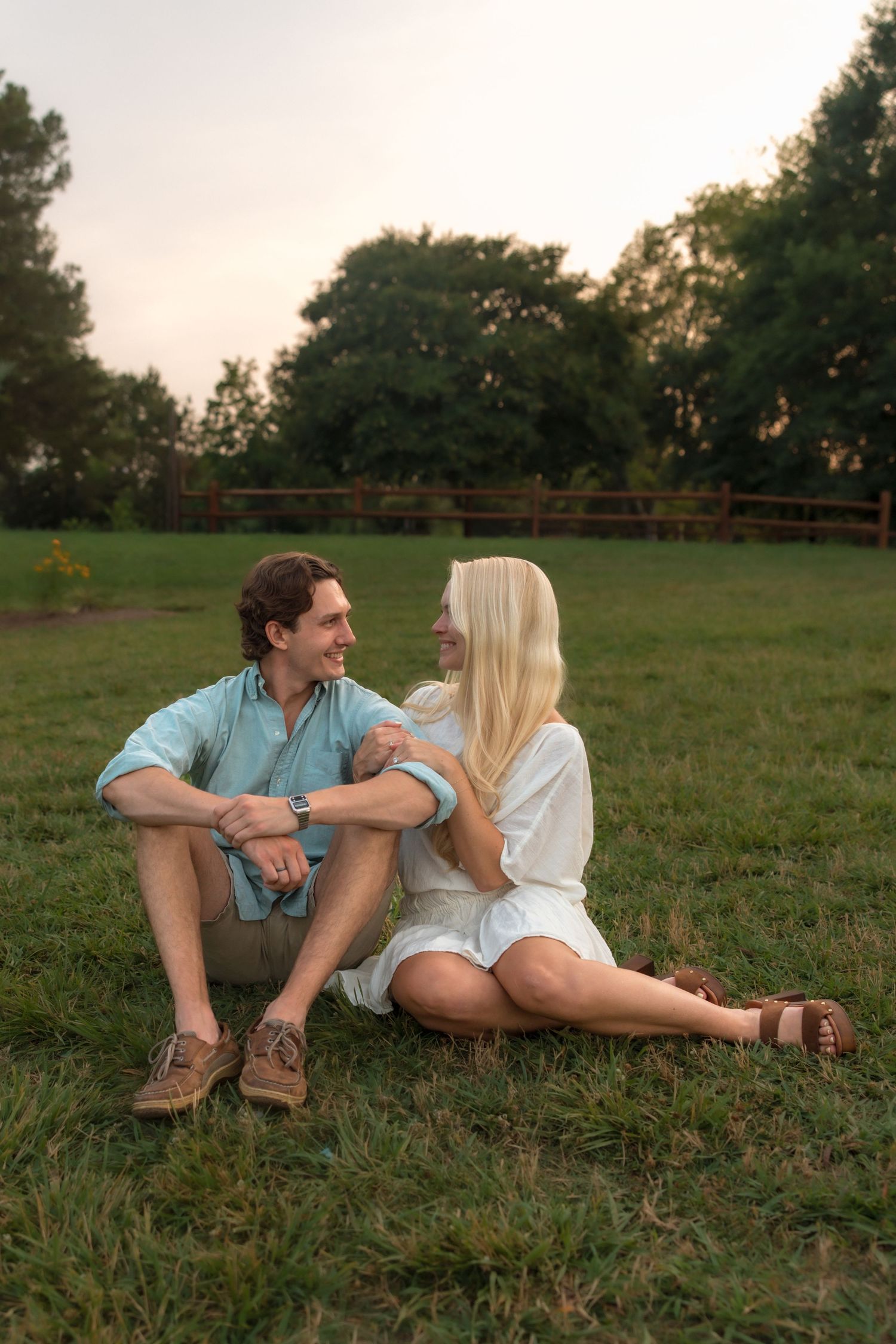 Romantic couple sitting on green grass in park, gazing at each other and smiling at sunset.