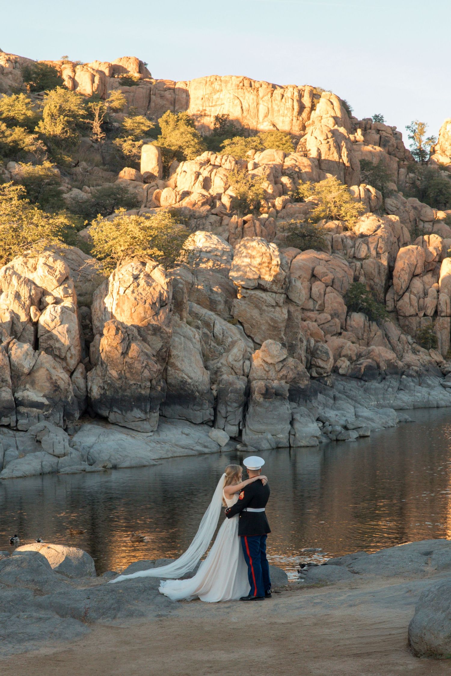 Bride and Marine groom embrace at waters edge before stunning golden-lit rocky canyon landscape.